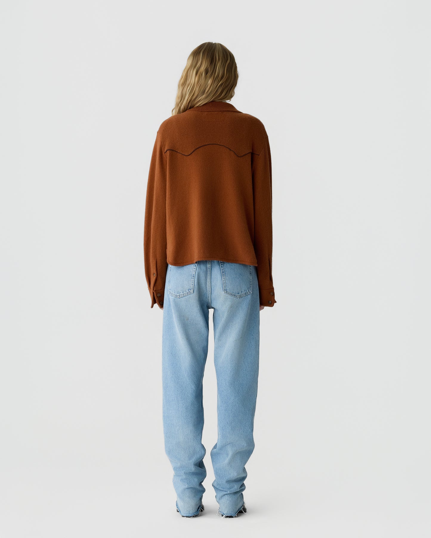 Woman with long blonde hair wearing a rust-colored sweater and light blue jeans, standing facing away against a plain light background.