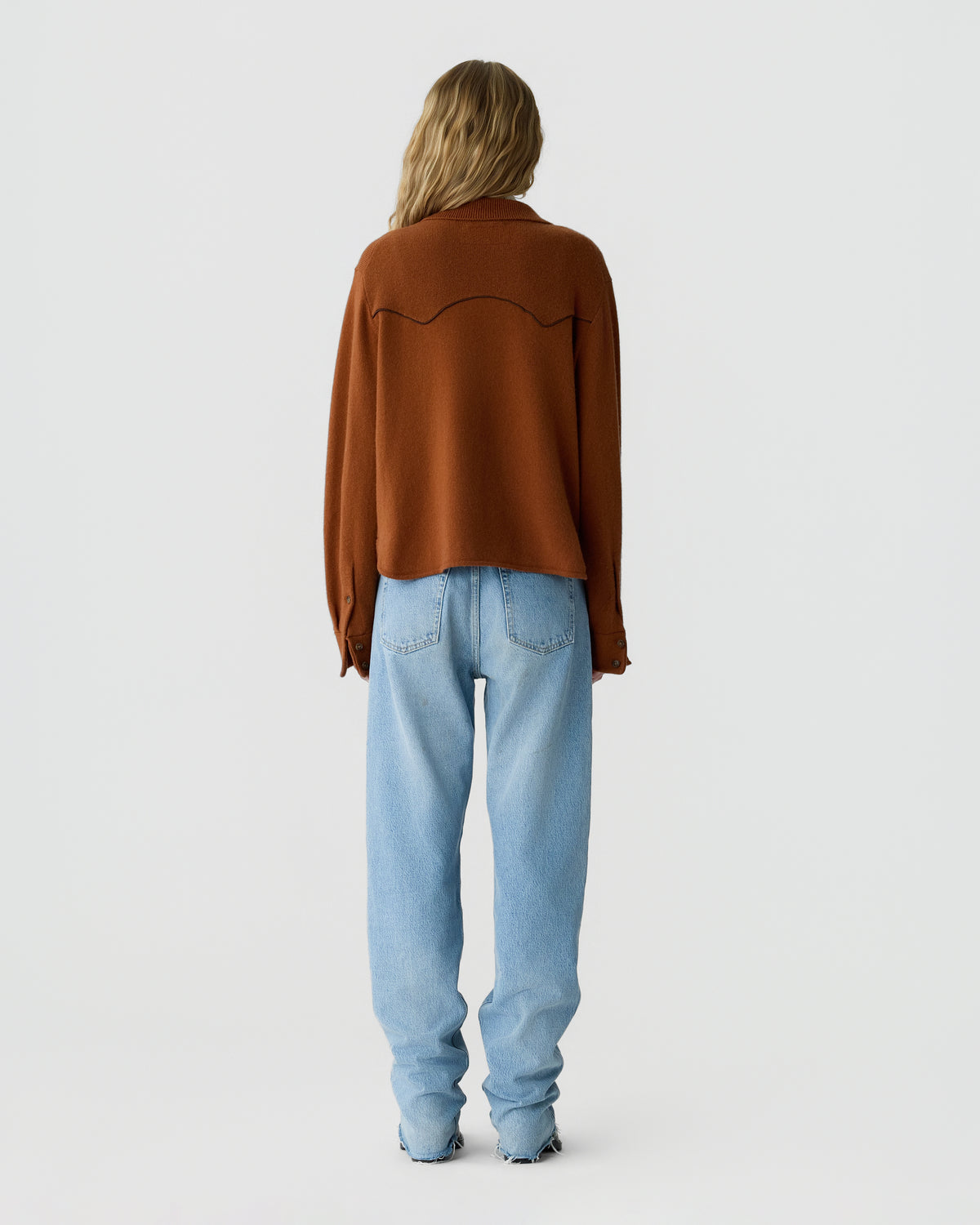 Back view of a woman wearing a rust brown long sleeve sweater with curved seam detailing and relaxed light blue jeans against a plain light gray background