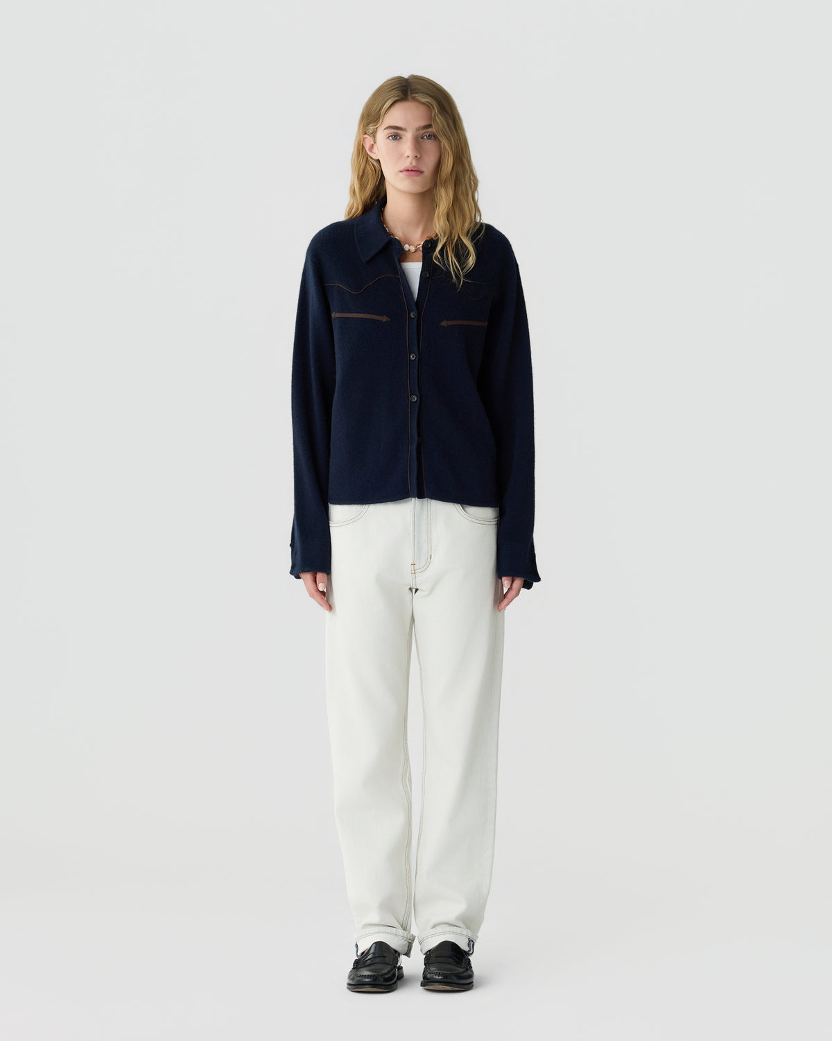 Young woman with long blonde hair wearing a dark navy button-up cardigan, white T-shirt, loose white pants, and black shoes, standing against a plain light background.