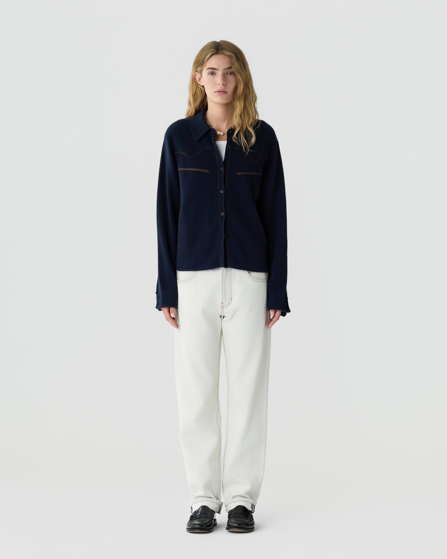 Young woman with long blonde hair wearing a dark navy button-up cardigan, white T-shirt, loose white pants, and black shoes, standing against a plain light background.