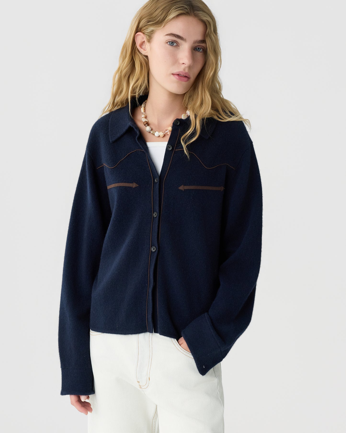 Young woman with long blonde hair wearing a navy blue button-up cashmere cardigan over a white top and white pants, accessorized with a chunky necklace, standing against a plain light grey background.