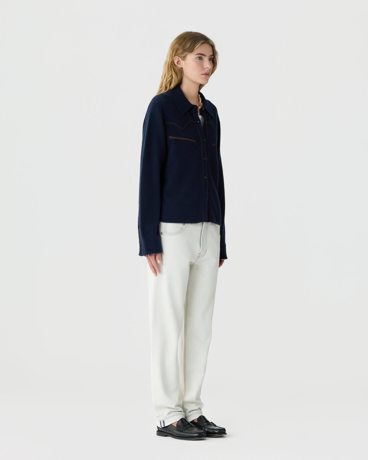 Young woman with long blonde hair wearing a navy blue button-up cashmere cardigan, white pants, and black loafers standing against a plain white background.