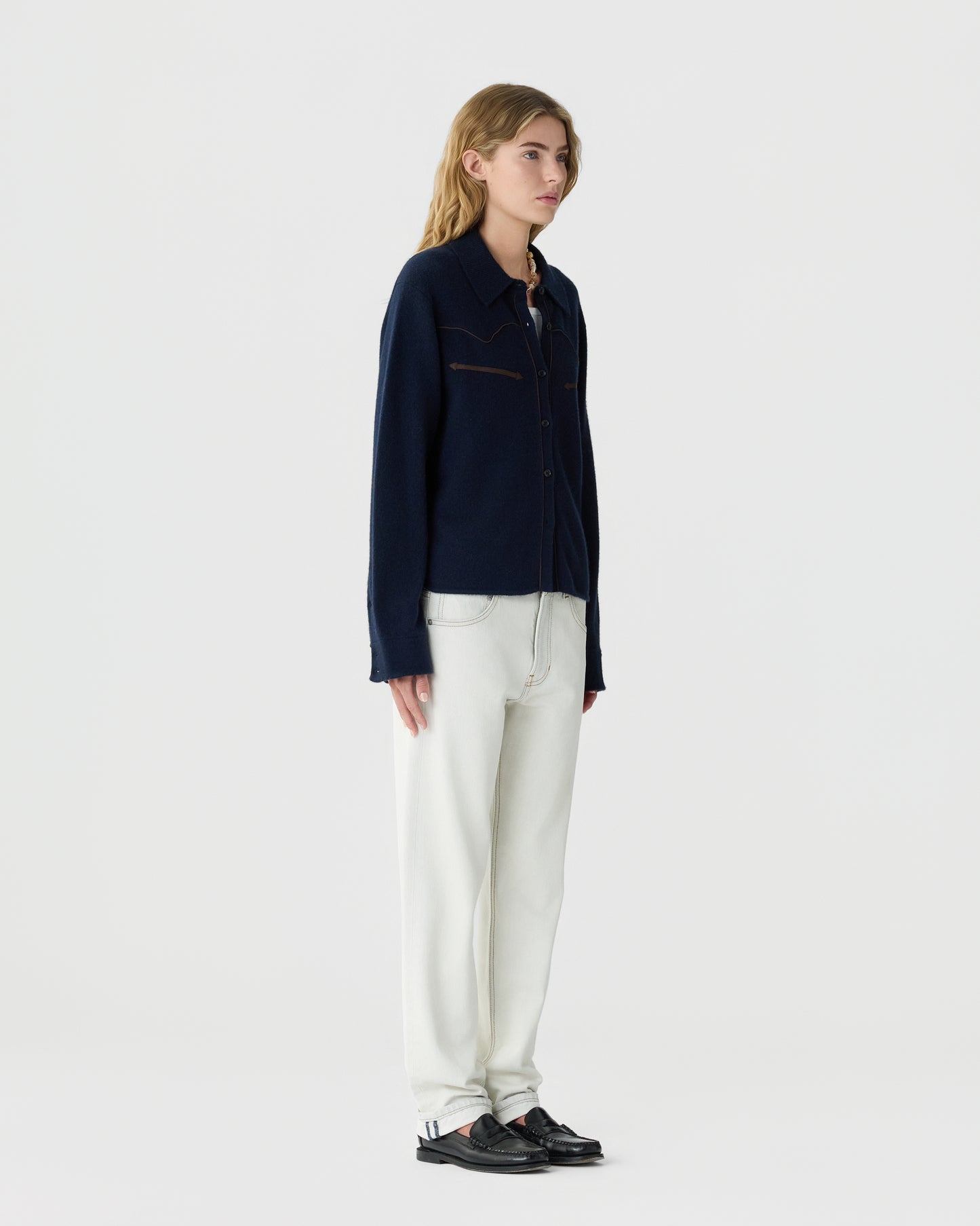 Young woman with long blonde hair wearing a navy blue button-up cashmere cardigan, white pants, and black loafers standing against a plain white background.