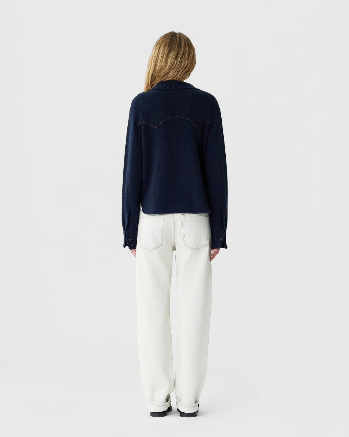 Woman with long blonde hair wearing a navy blue long-sleeve top and high-waisted white pants standing facing away against a plain white background.