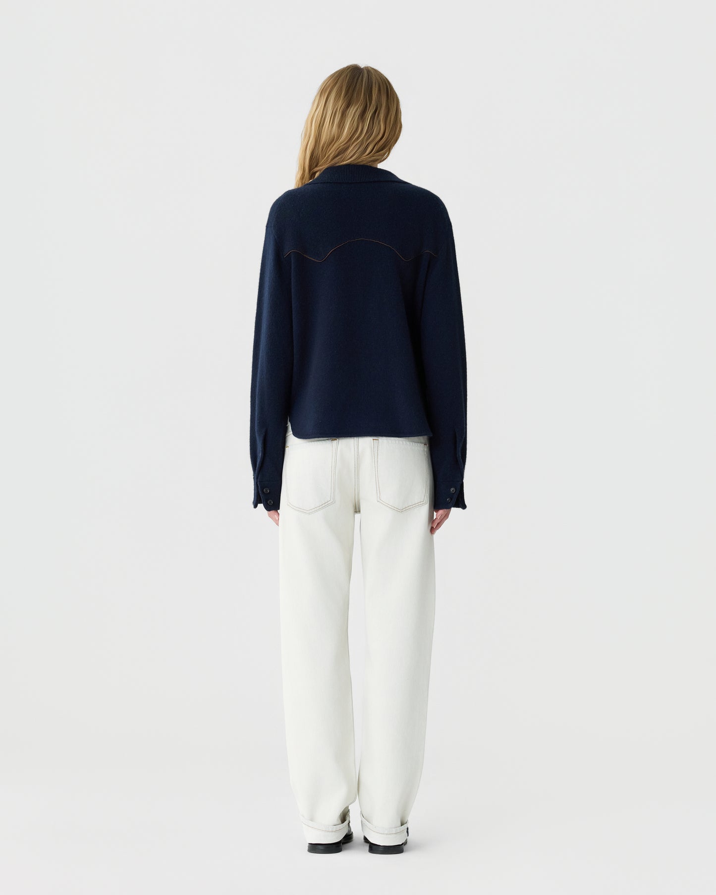 Woman with long blonde hair wearing a navy blue long-sleeve top and high-waisted white pants standing facing away against a plain white background.