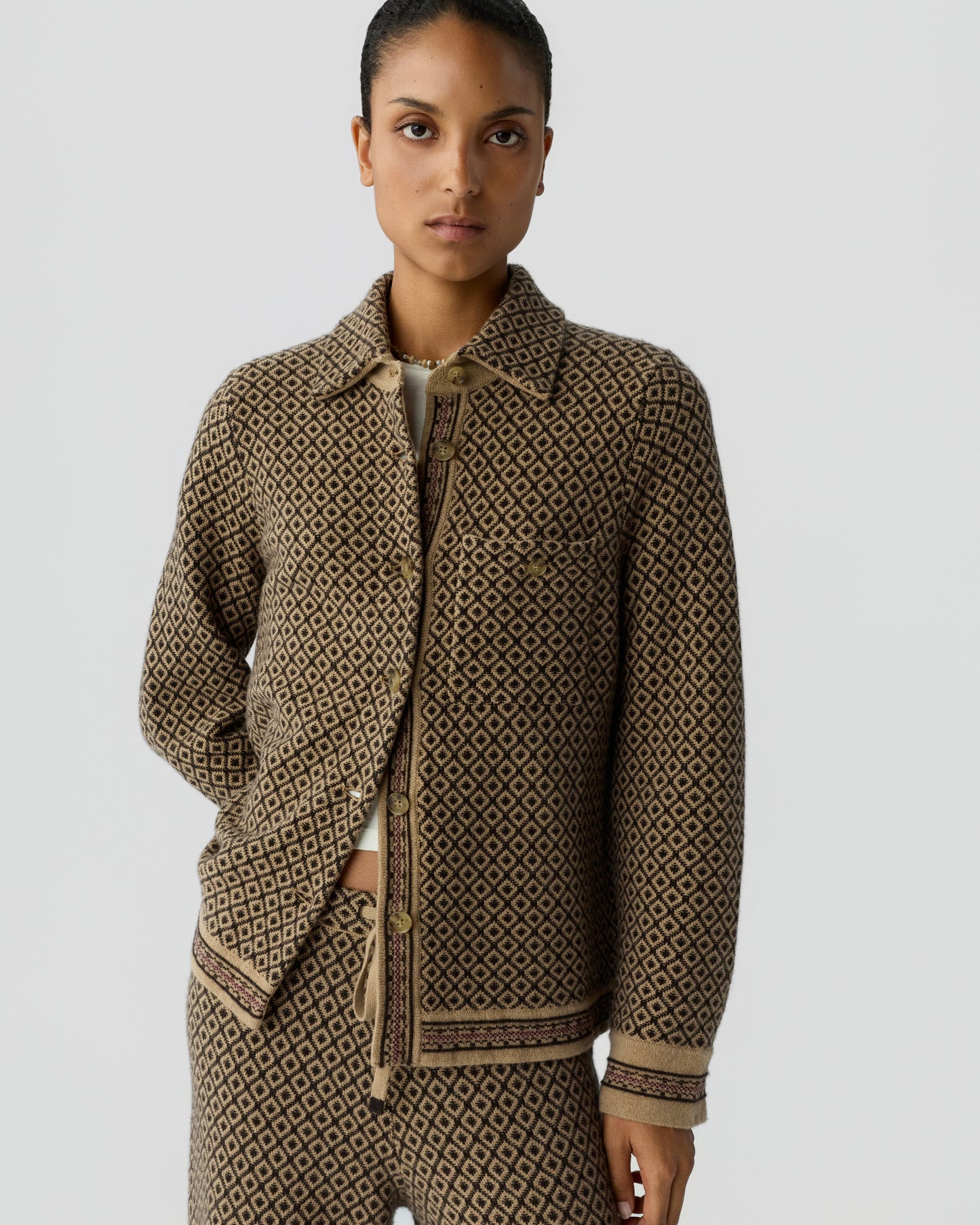 Woman wearing a brown and black patterned jacket and matching pants with a geometric diamond design, standing against a plain light gray background.