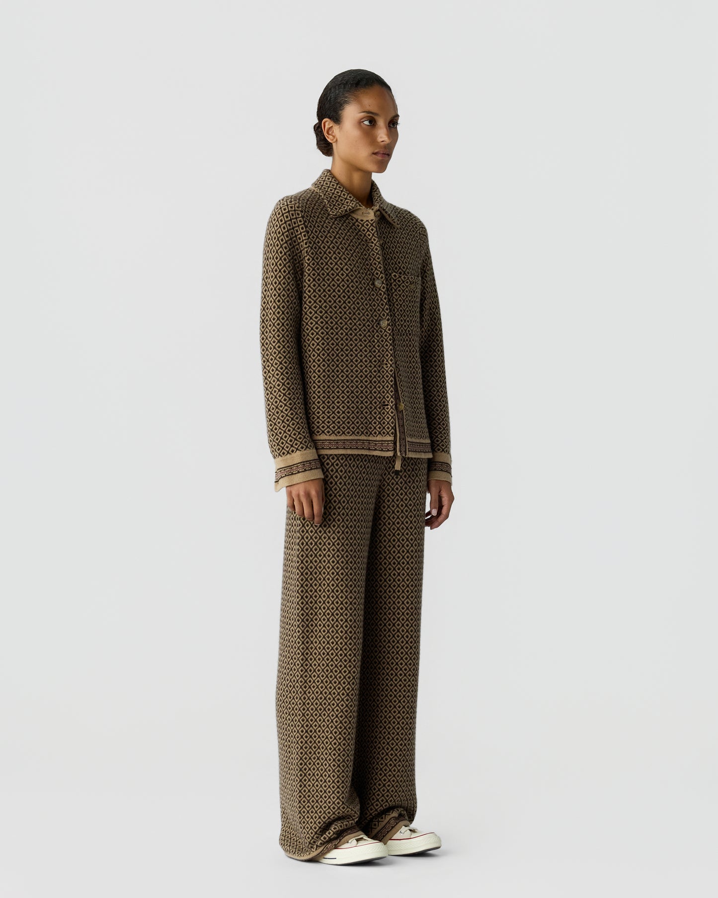Woman standing wearing a matching brown and black patterned knit shirt and wide-leg pants paired with white sneakers against a plain light background.