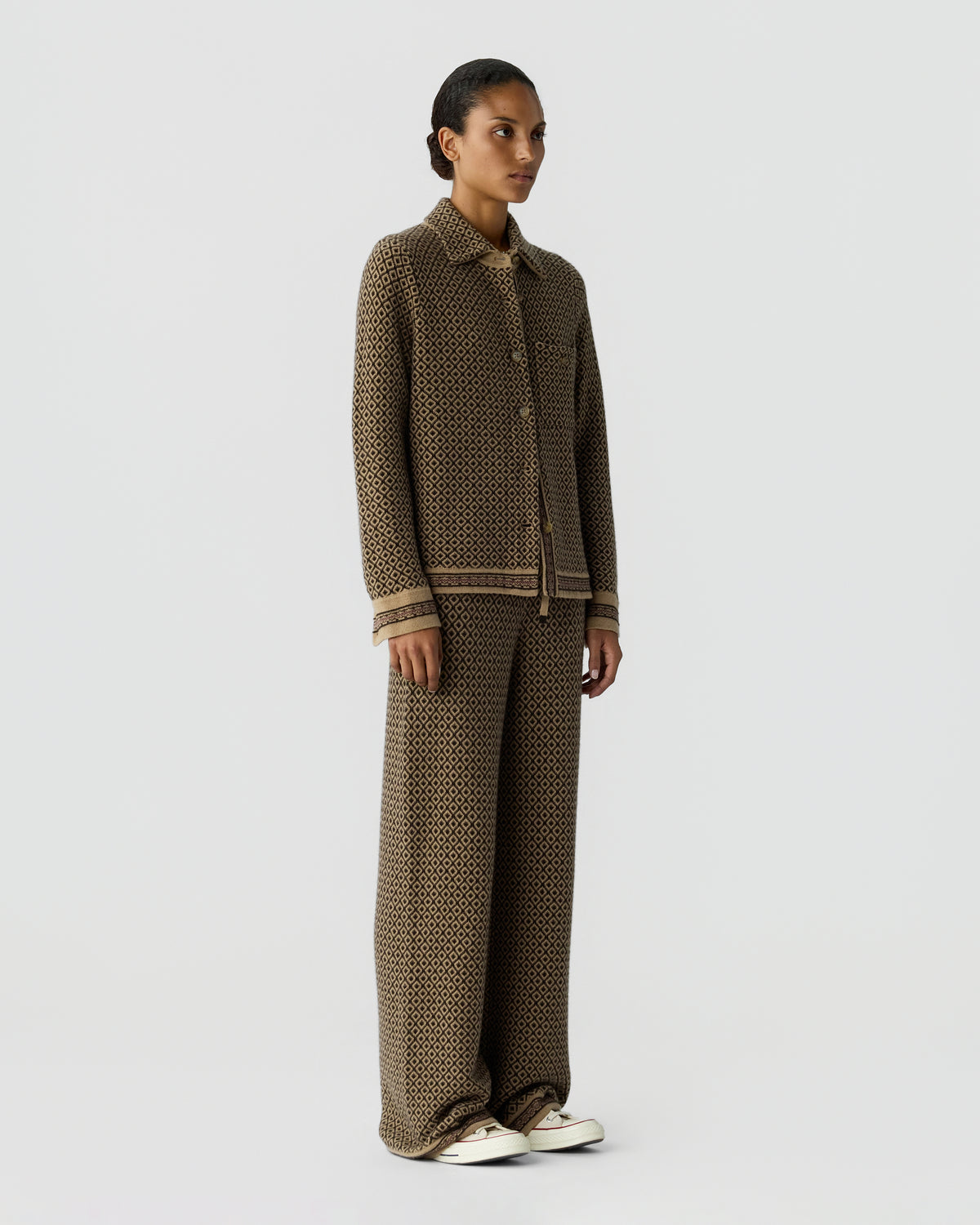 Model wearing a brown cashmere knit jacket and matching wide leg trousers with white sneakers against a light gray background