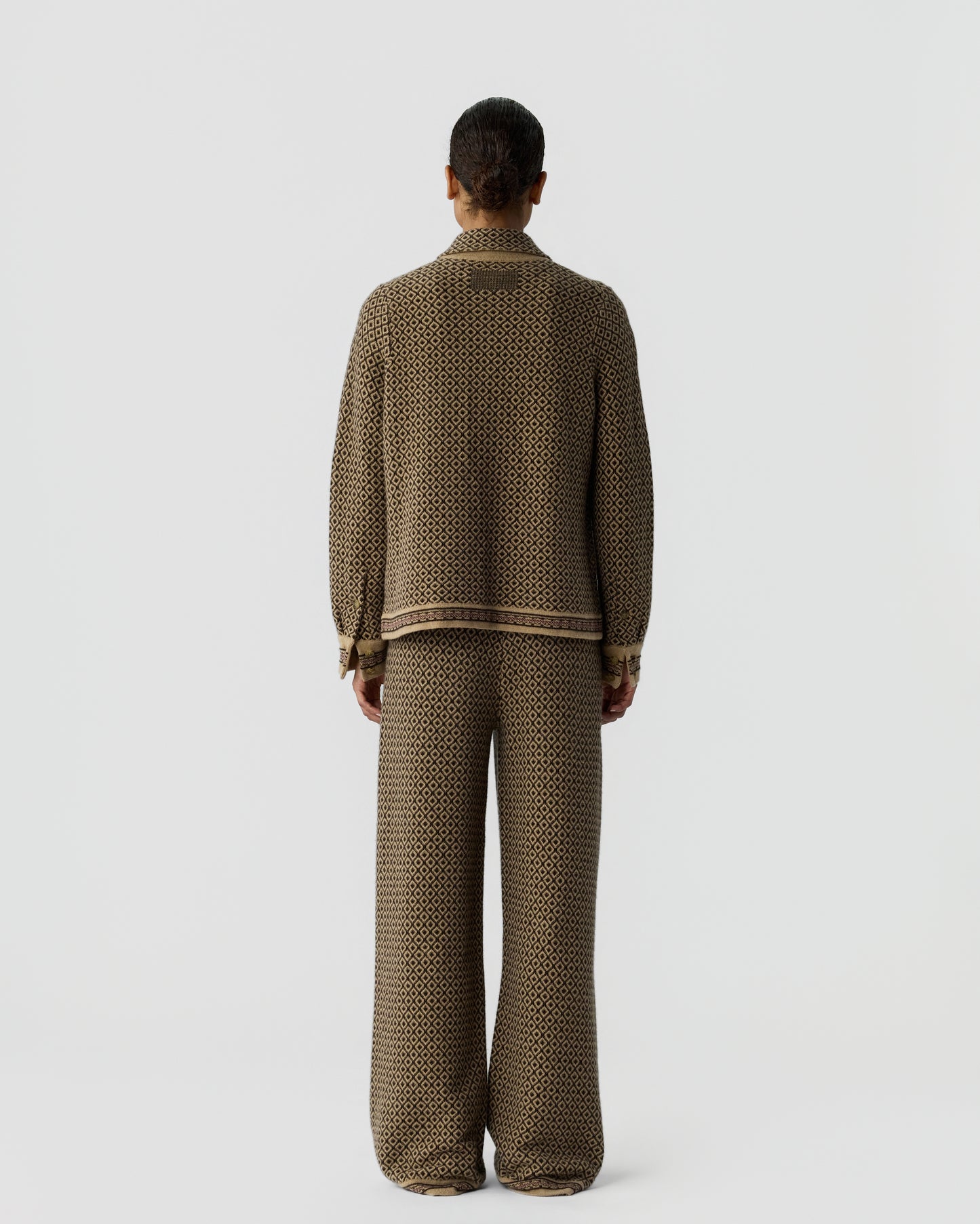 Person seen from behind wearing a matching brown and black patterned knit sweater and wide-leg pants standing against a plain light gray background.