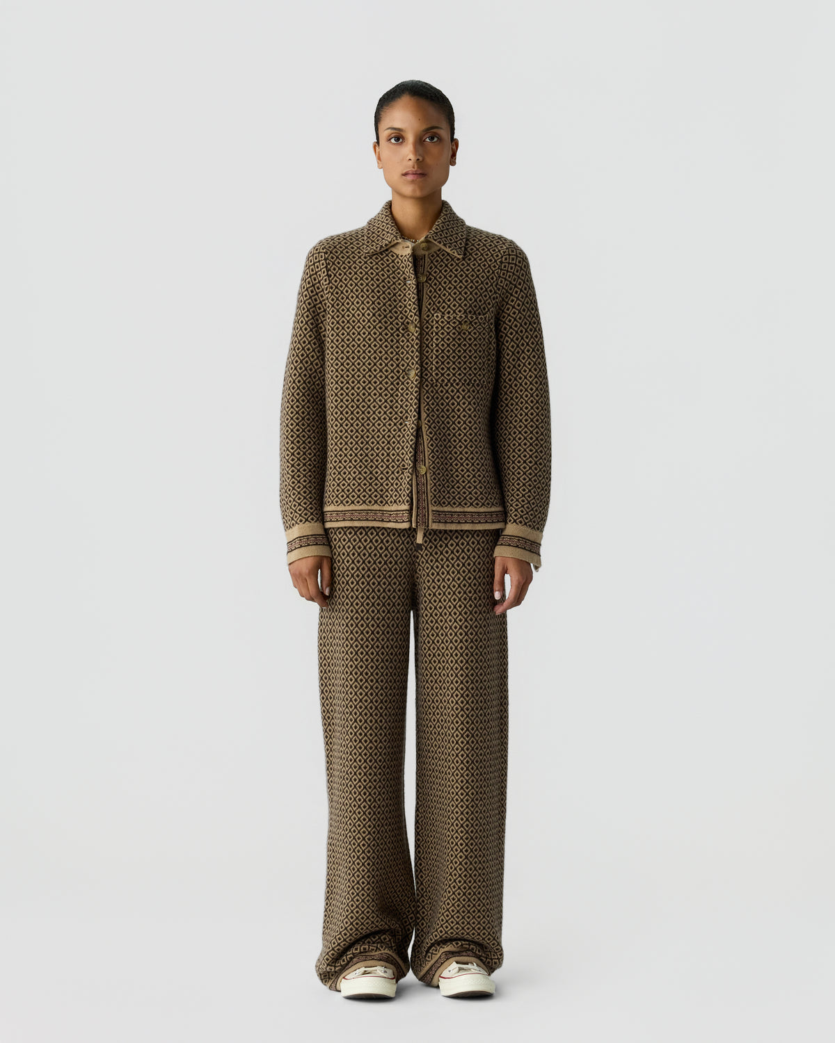 Woman wearing a matching brown patterned knit jacket and wide-leg pants standing against a plain light gray background with neutral expression and white shoes.