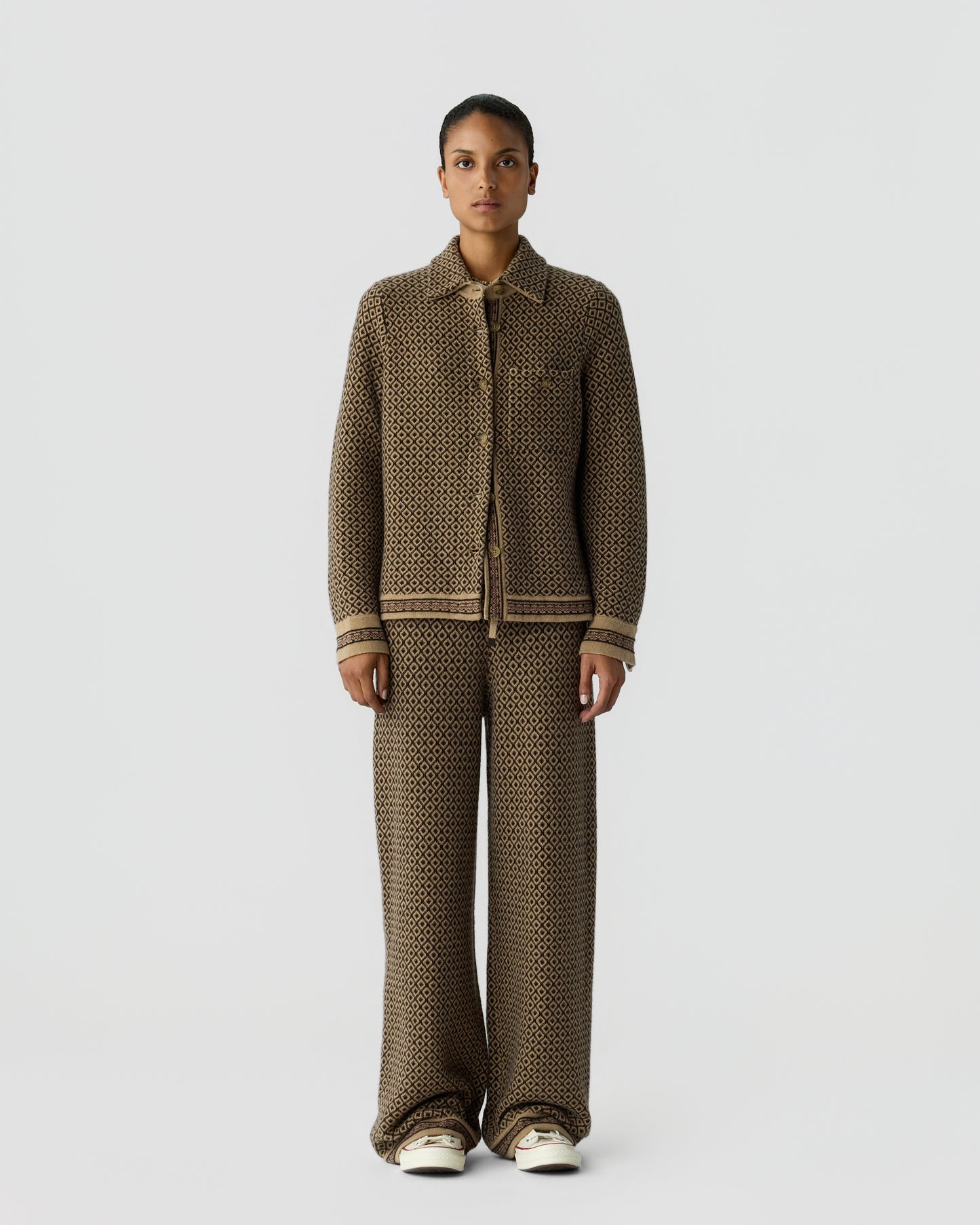 Woman wearing a matching brown patterned knit jacket and wide-leg pants standing against a plain light gray background with neutral expression and white shoes.