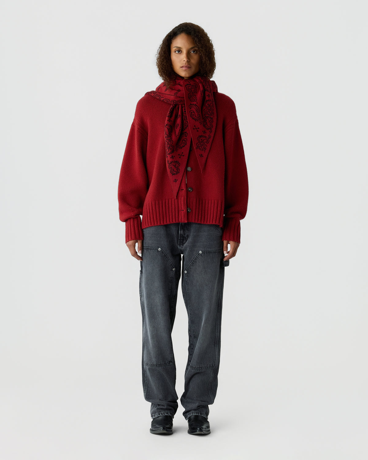 Model wearing a deep red buttoned cardigan with a coordinating patterned scarf, dark gray jeans, and black shoes against a light background.