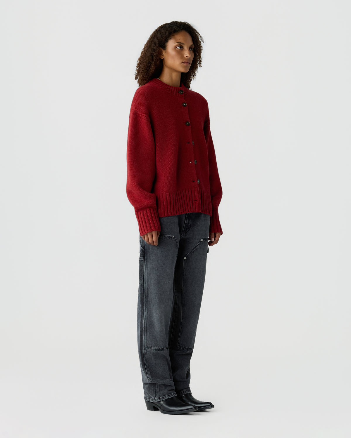 Woman standing in a studio wearing a deep red button front cardigan, dark gray jeans, and black shoes against a light gray background