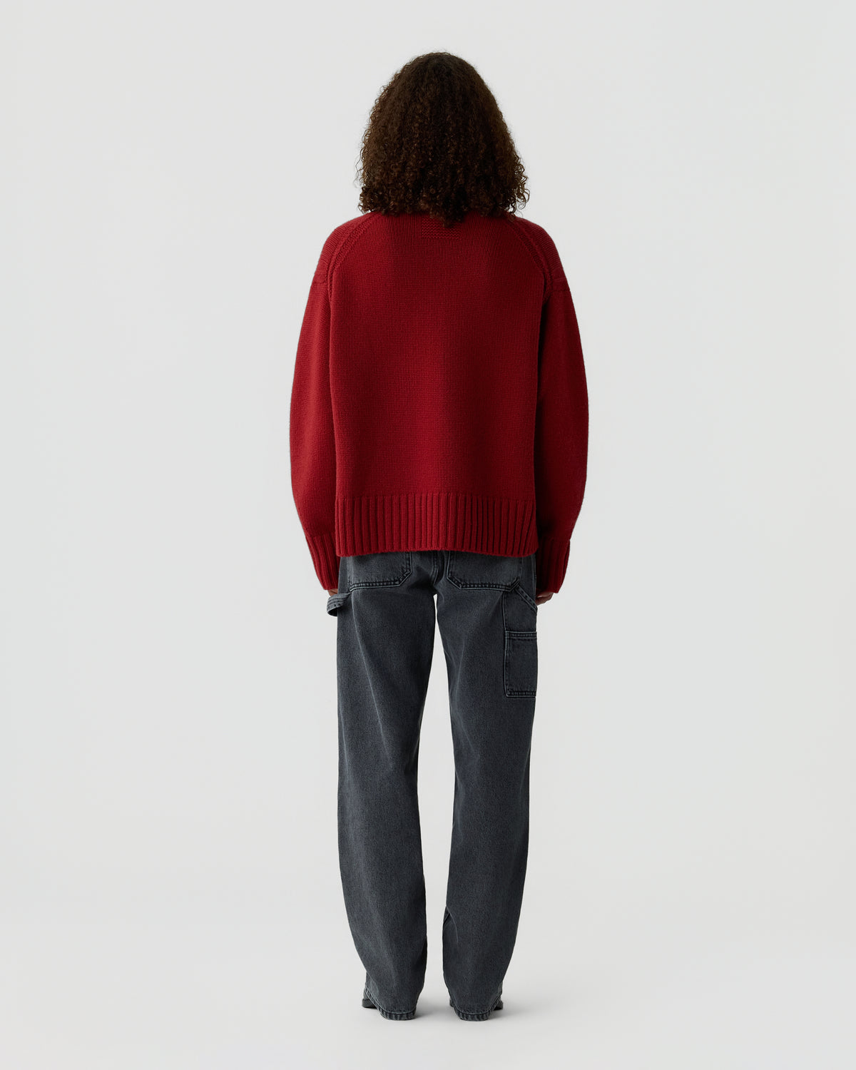 Person viewed from behind wearing a deep red ribbed sweater with dark gray cargo-style trousers and shoulder-length curly hair against a light background