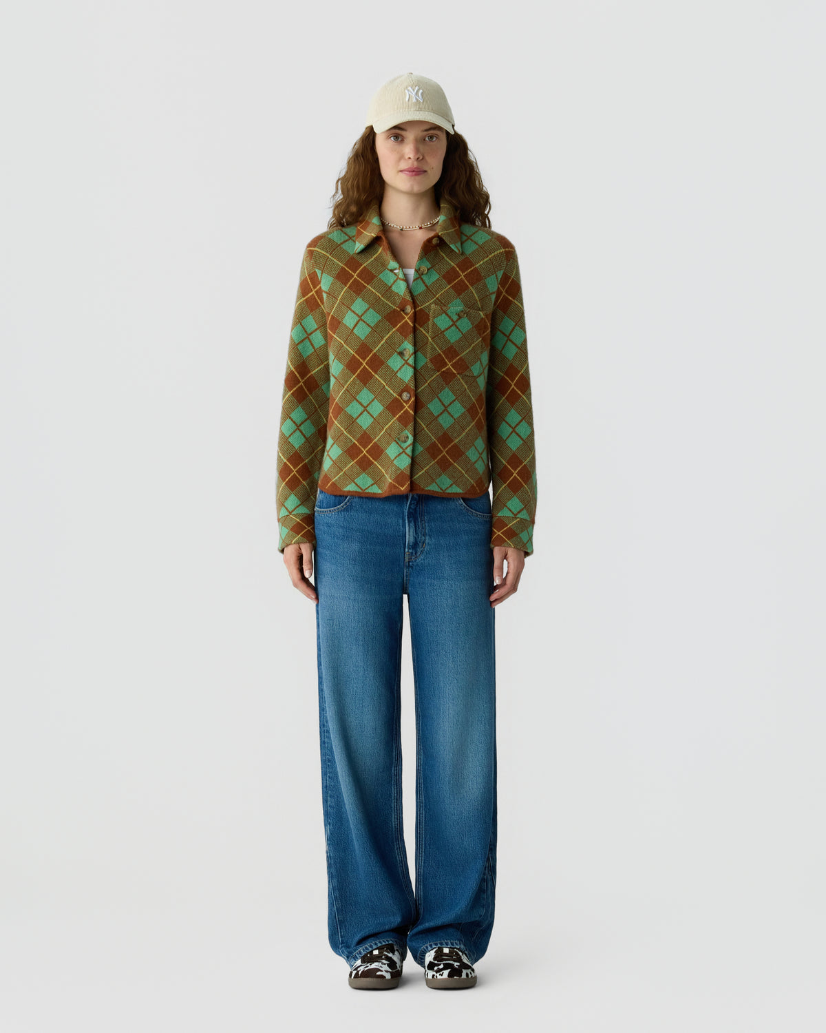 Model wearing a green and brown plaid button up shirt with blue wide leg jeans a beige baseball cap and sneakers against a light background