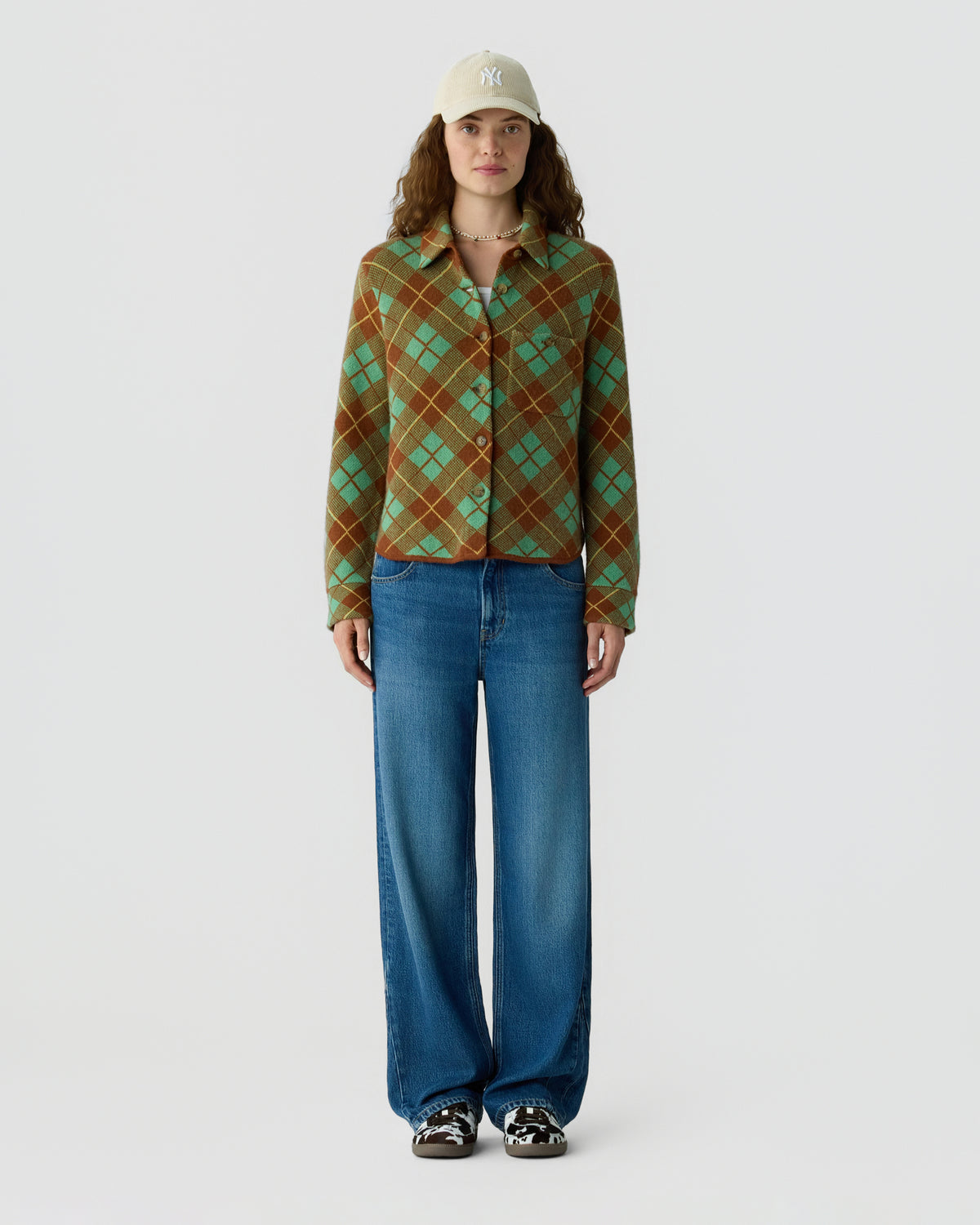 Woman wearing a green and brown argyle button up sweater with wide leg blue jeans sneakers and a beige cap against a light gray background
