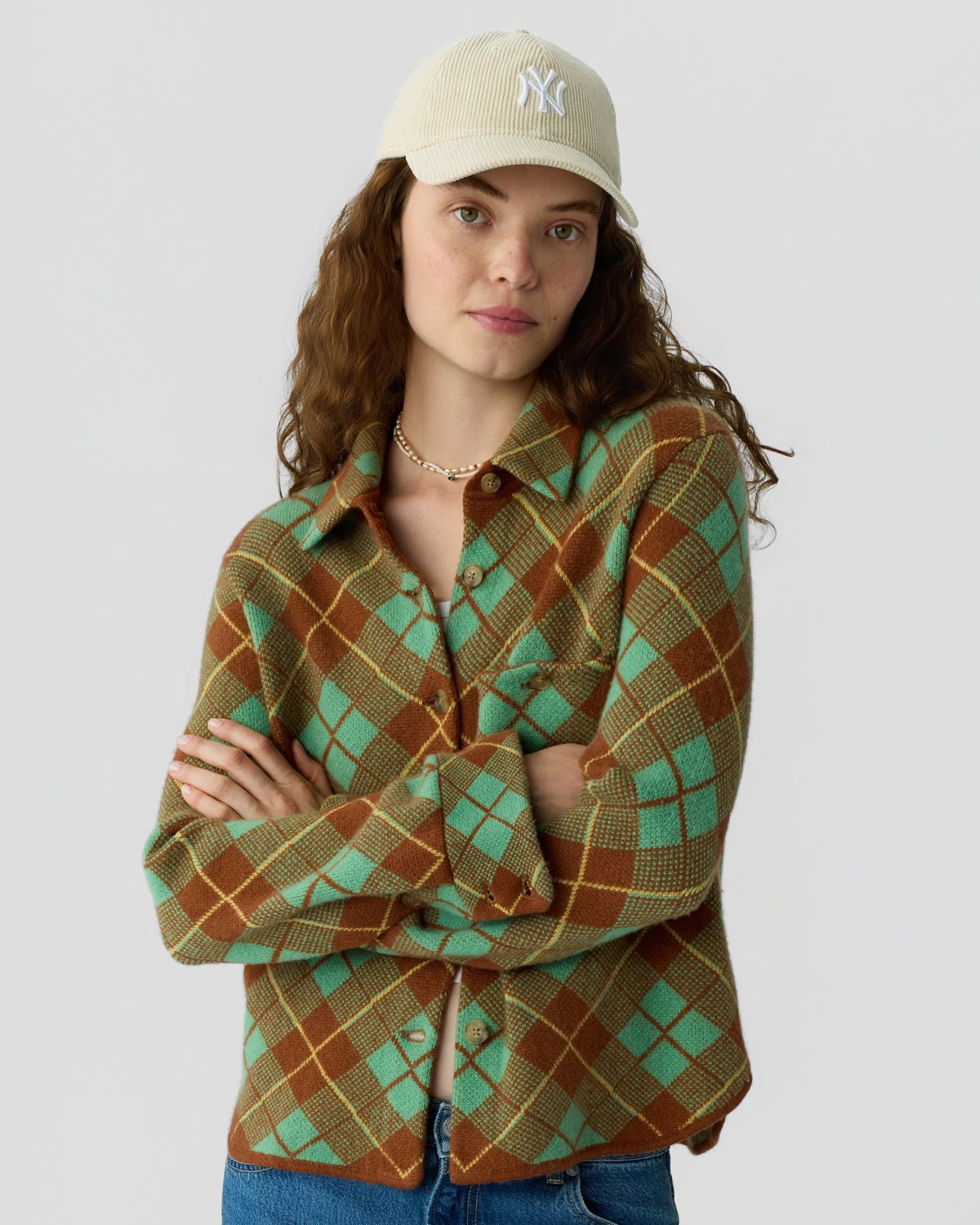 Woman wearing a green and brown argyle button front shirt with rolled cuffs and a beige baseball cap while standing against a light gray background