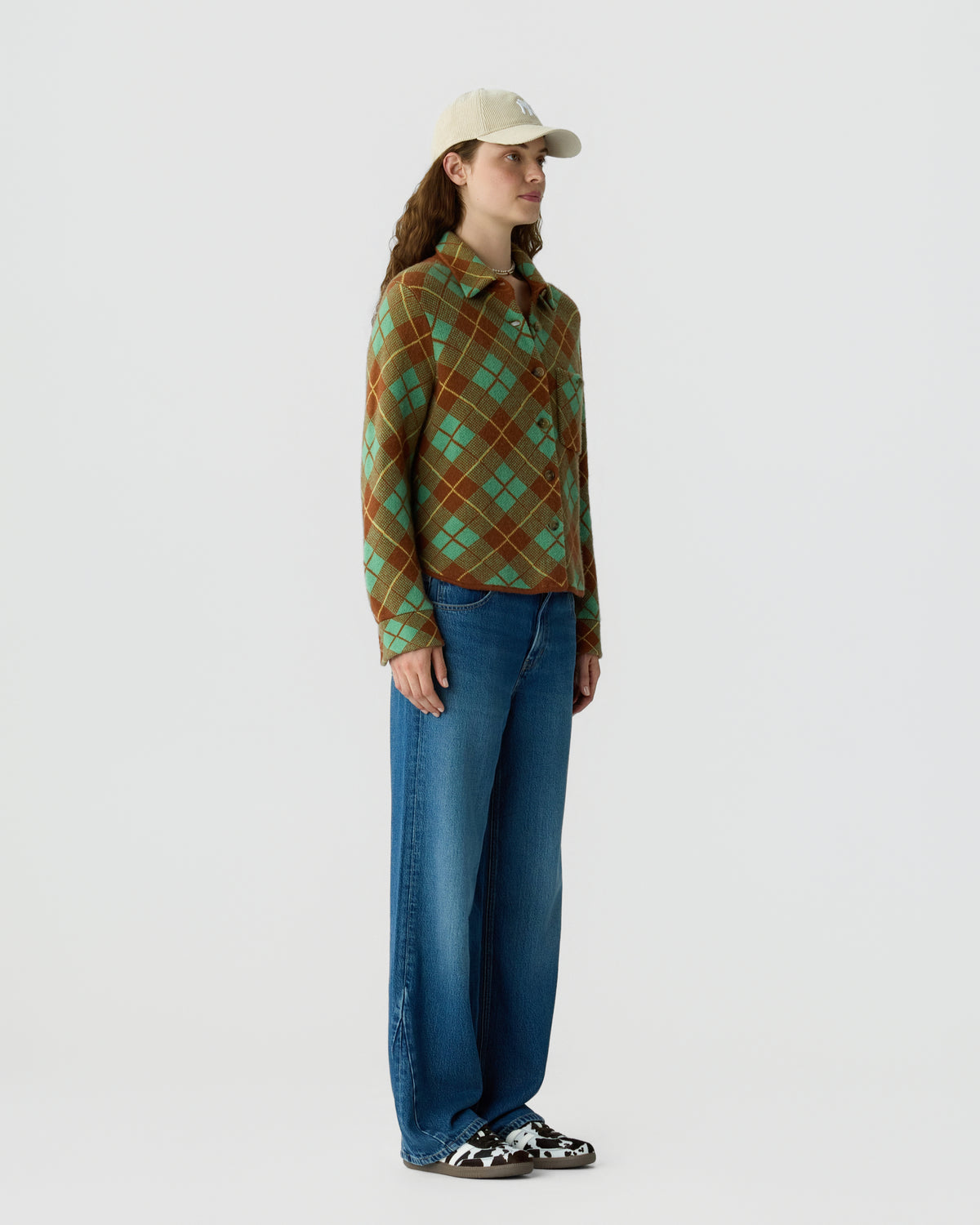 Woman wearing a brown and green plaid sweater with blue jeans a beige cap and sneakers against a light gray background
