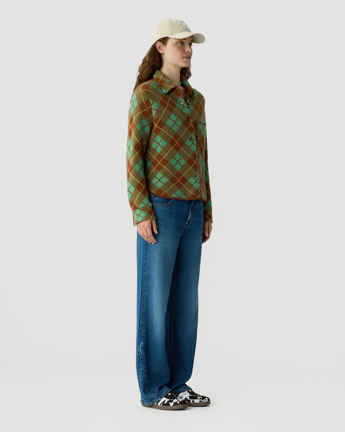 Model wearing a green and brown plaid cashmere shirt with wide leg blue jeans, a beige baseball cap, and black and white sneakers against a light background
