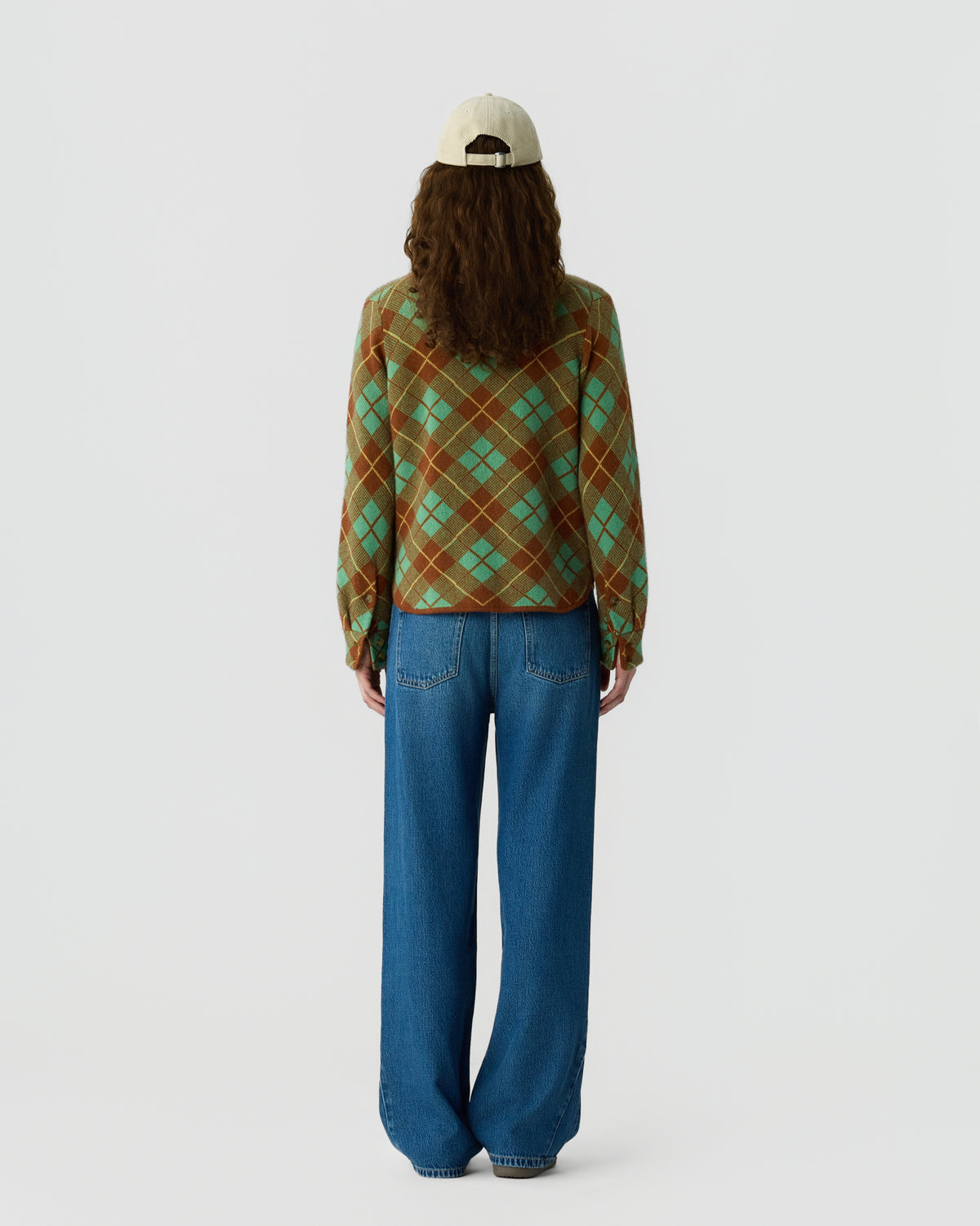 Rear view of a person wearing a brown and green argyle sweater, blue wide leg jeans, and a white baseball cap against a light gray background