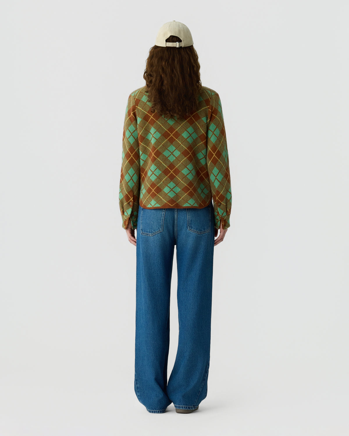 Back view of a person wearing a green and brown argyle sweater with blue jeans and a cream baseball cap against a light gray background