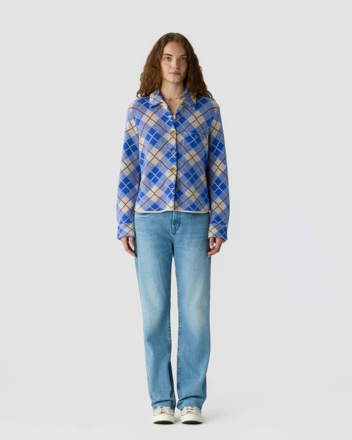 Person standing against a light background wearing a blue and white plaid button front sweater with light blue jeans and white sneakers