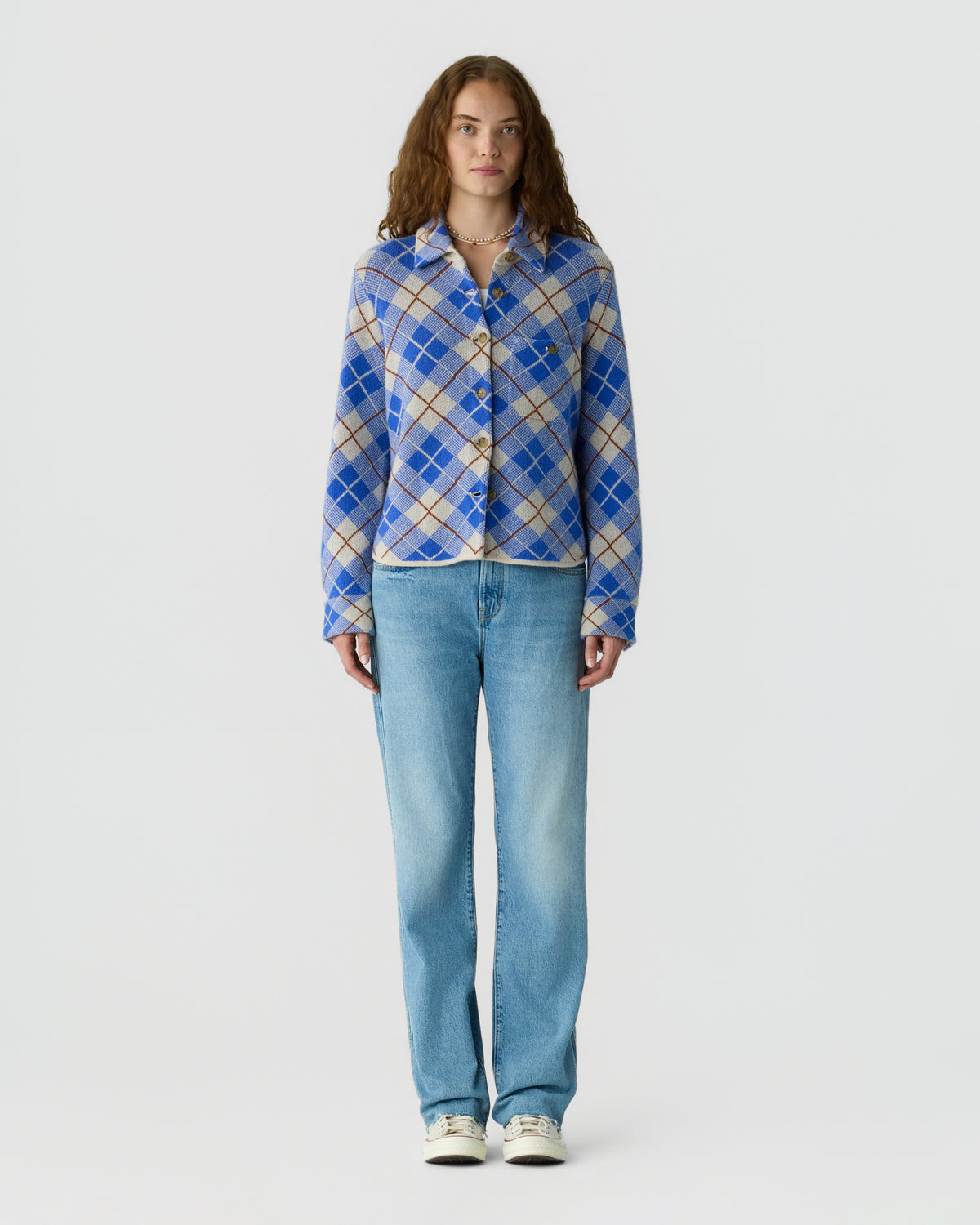 Full length studio portrait of a woman wearing a blue and white plaid button up jacket with light blue jeans and white sneakers against a pale gray background