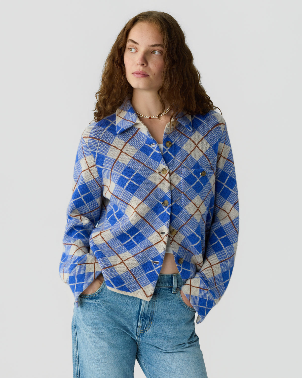 Woman with wavy brown hair wearing a blue and cream plaid button front cardigan with jeans against a light gray background