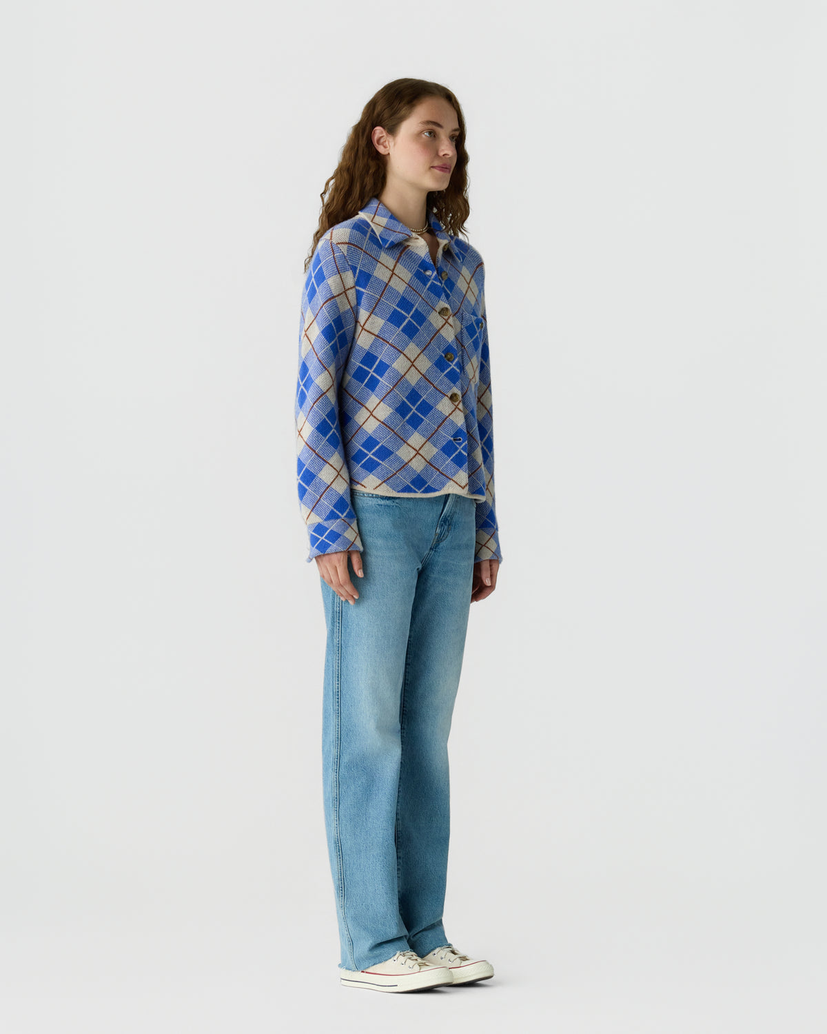 Woman wearing a blue plaid knit sweater, light blue jeans, and white sneakers while standing against a plain light gray background.
