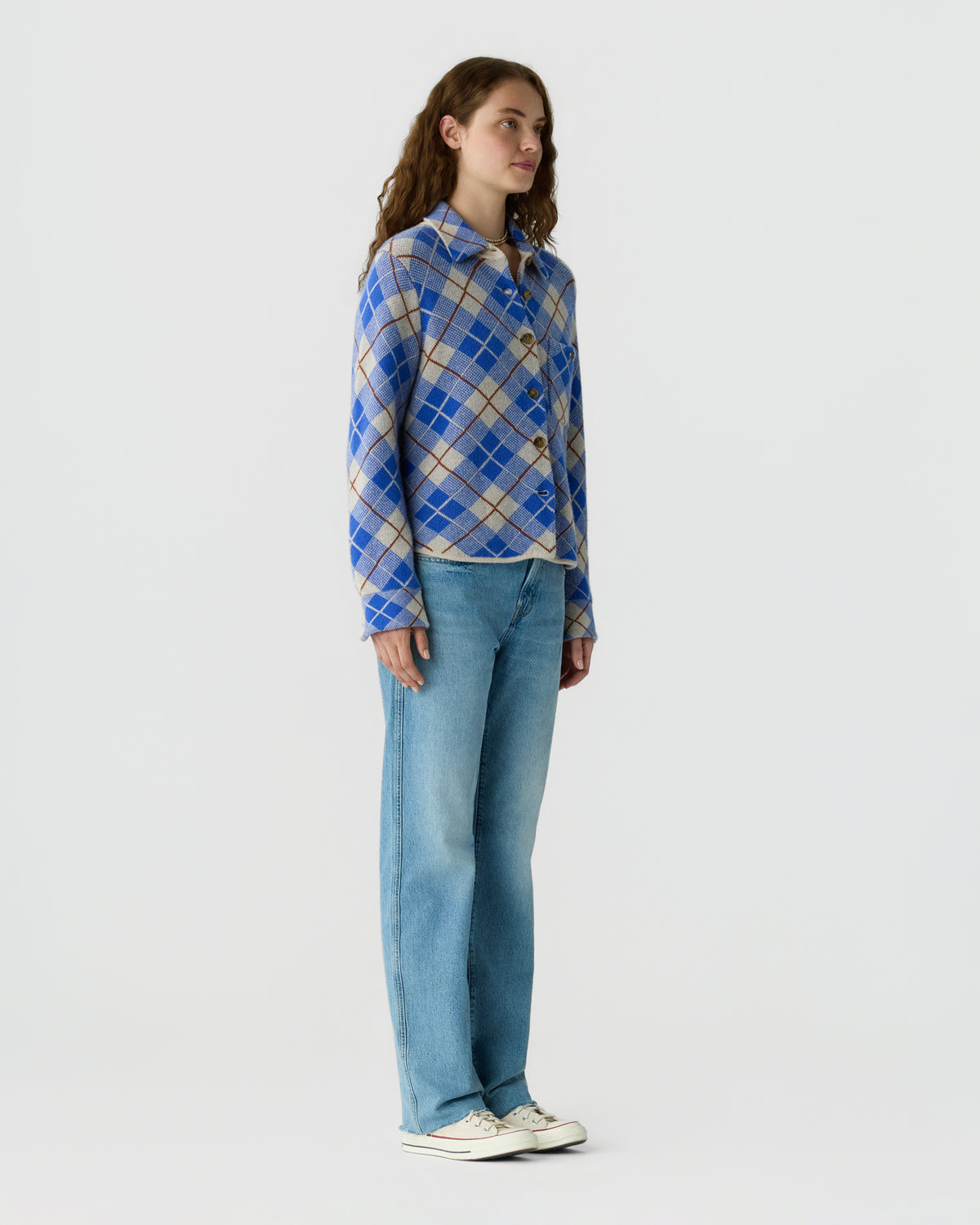 Woman standing in a blue and beige plaid sweater with light blue jeans and white sneakers against a plain light gray background