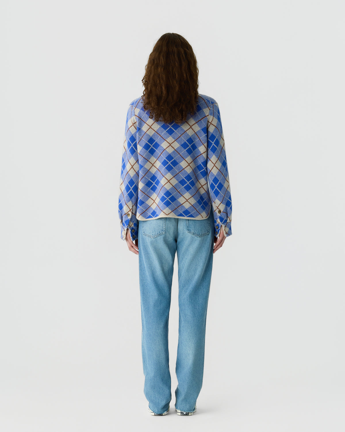 Back view of a woman wearing a blue and cream argyle sweater with loose sleeves and light blue jeans against a plain white background