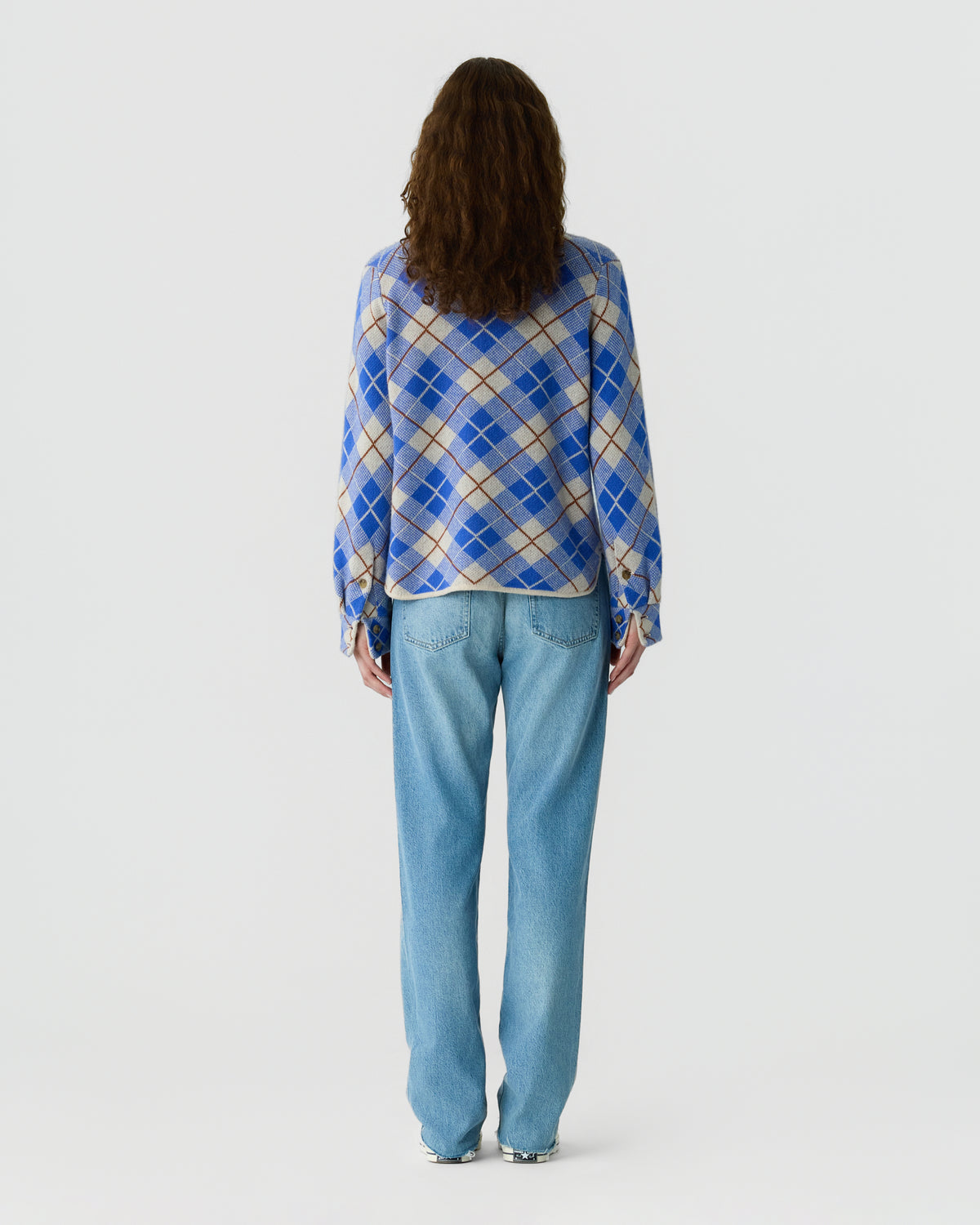 Rear view of a woman wearing a blue and cream argyle cardigan with light blue jeans against a plain white background
