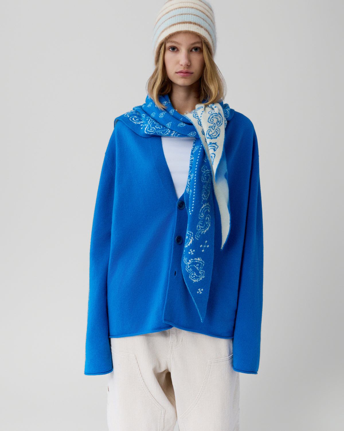 Young woman wearing a vibrant blue buttoned cardigan layered over a white top, paired with cream-colored pants, a blue patterned scarf, and a cream and blue striped beanie against a plain background.