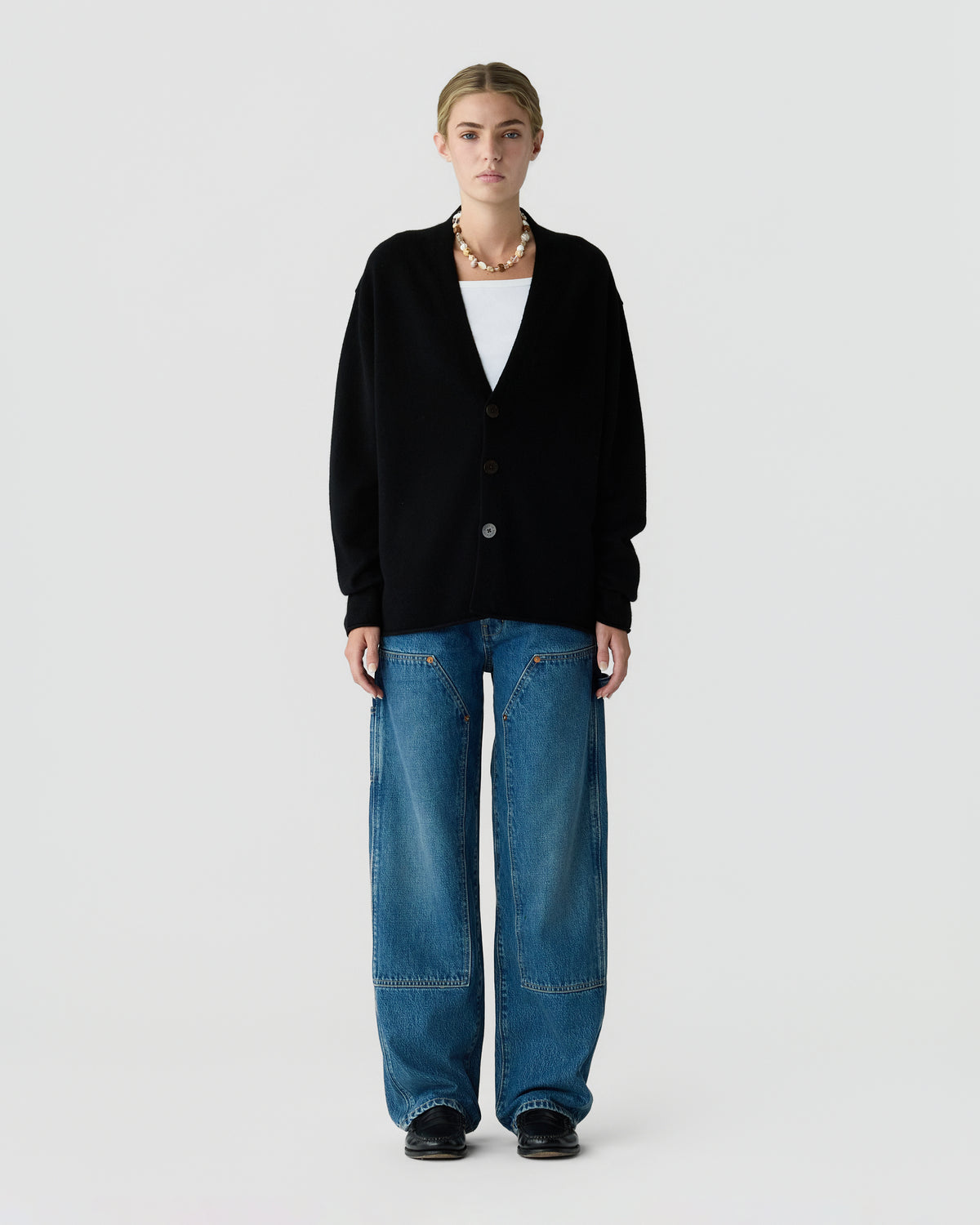 Person wearing a black button front cardigan over a white top, wide leg blue jeans, and black shoes against a light gray background