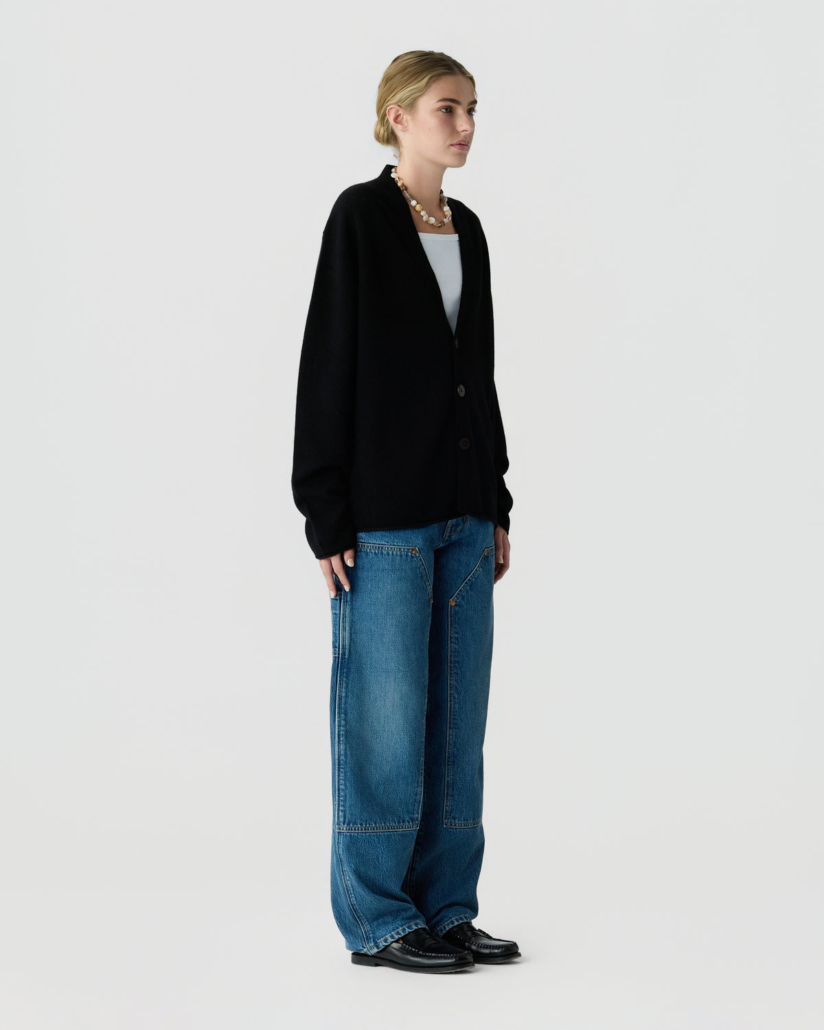Model wearing a black button front cardigan over a white top, wide leg blue jeans, a pearl necklace, and black loafers against a light background
