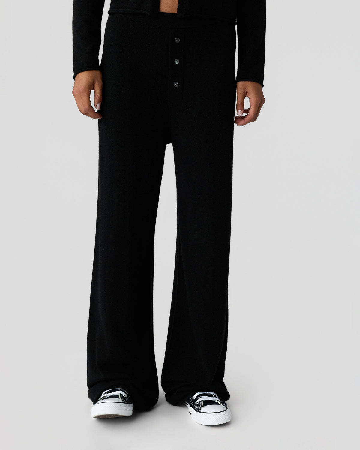 Person wearing black wide-leg trousers with a button fly and black long-sleeve top, paired with black and white sneakers against a light background.
