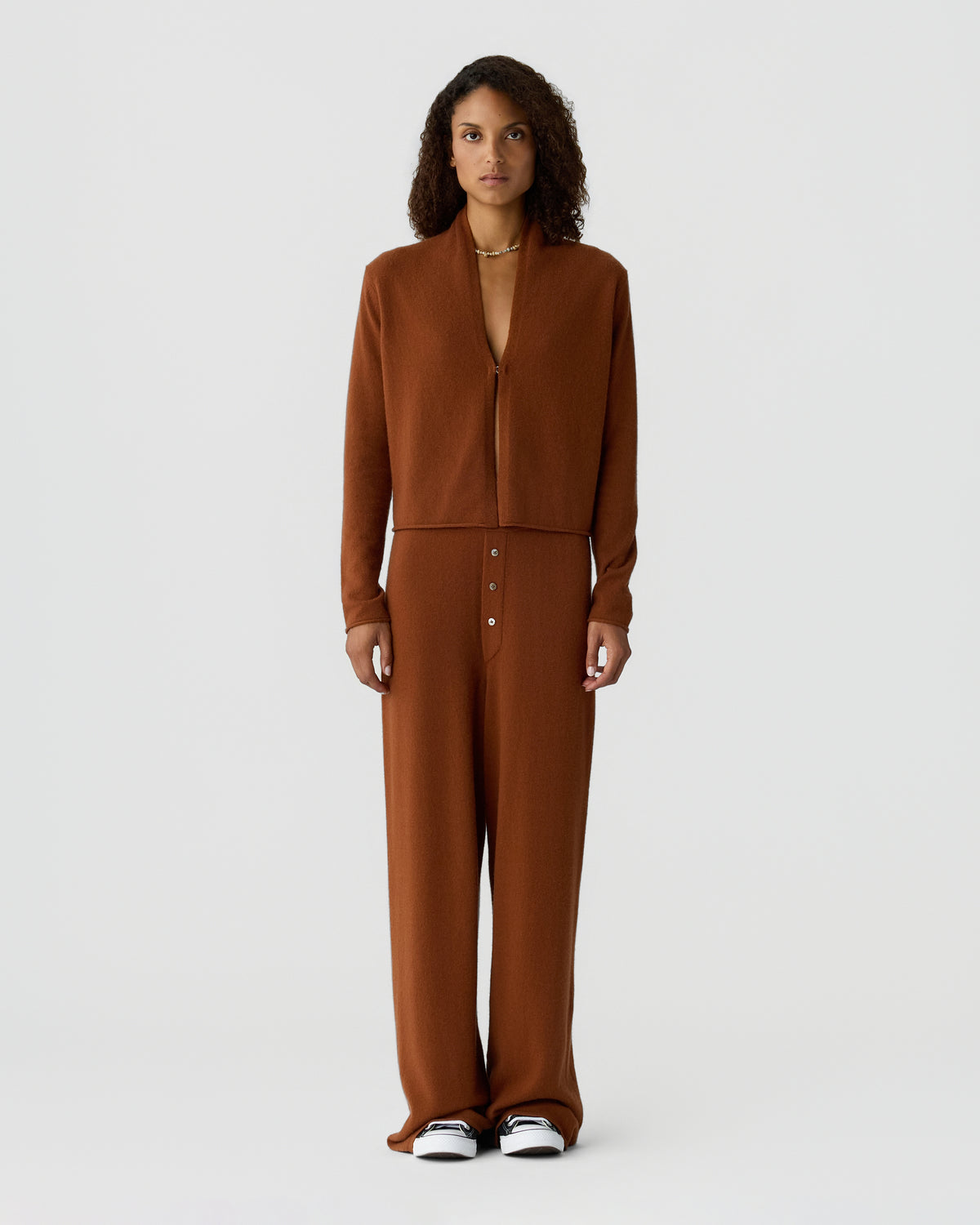 Full-length view of a woman wearing a brown zip-front jacket with matching high-waisted trousers and black and white sneakers against a light gray background