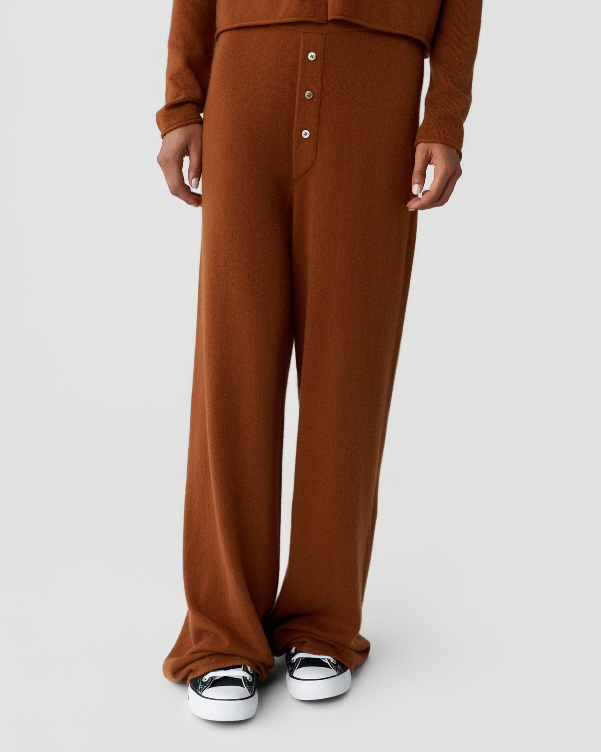 Lower half of a person wearing brown wide leg knit trousers with front buttons and white sneakers against a light gray background