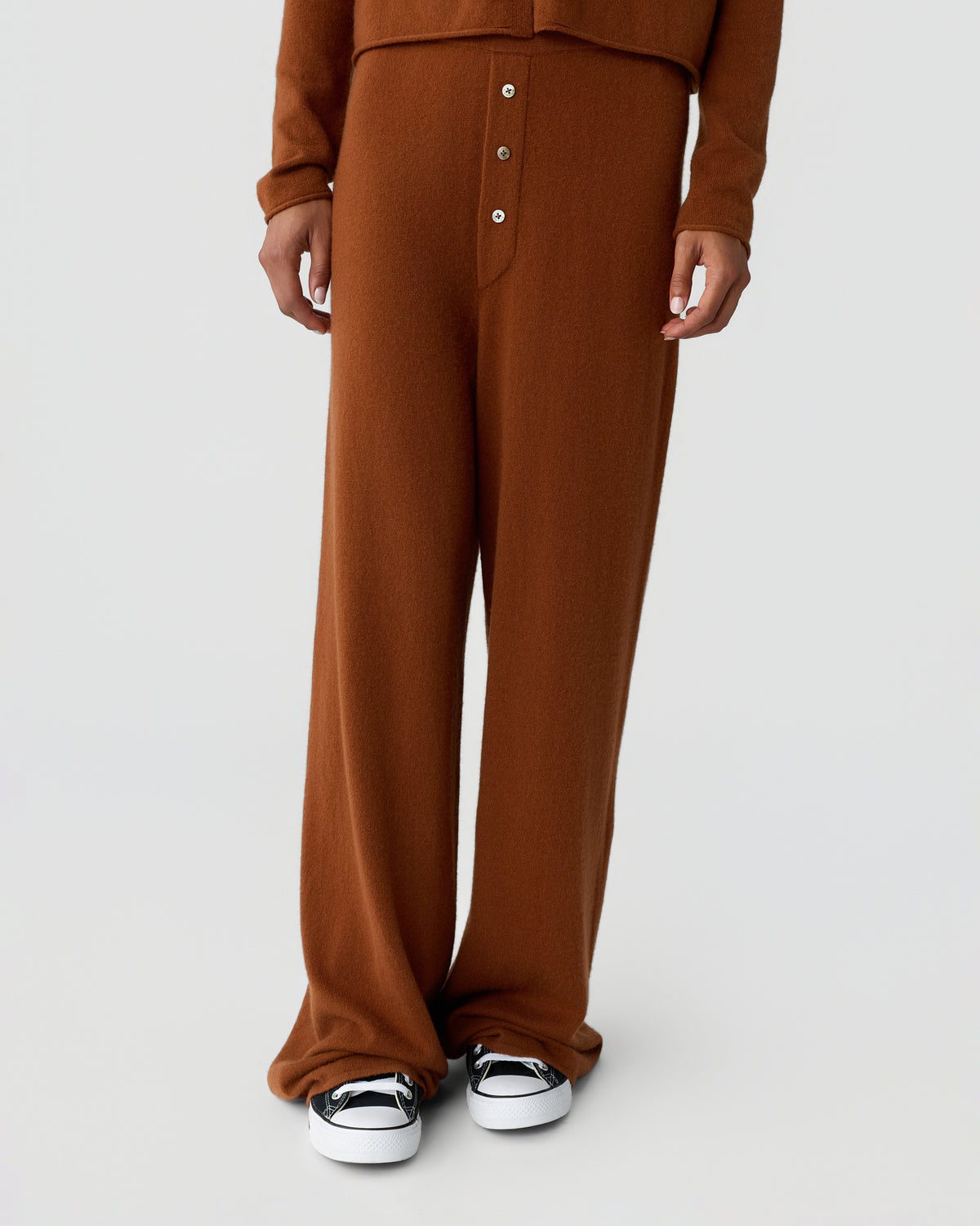 Model wearing a rust brown button front knit top and wide leg trousers with white and black sneakers against a light gray background