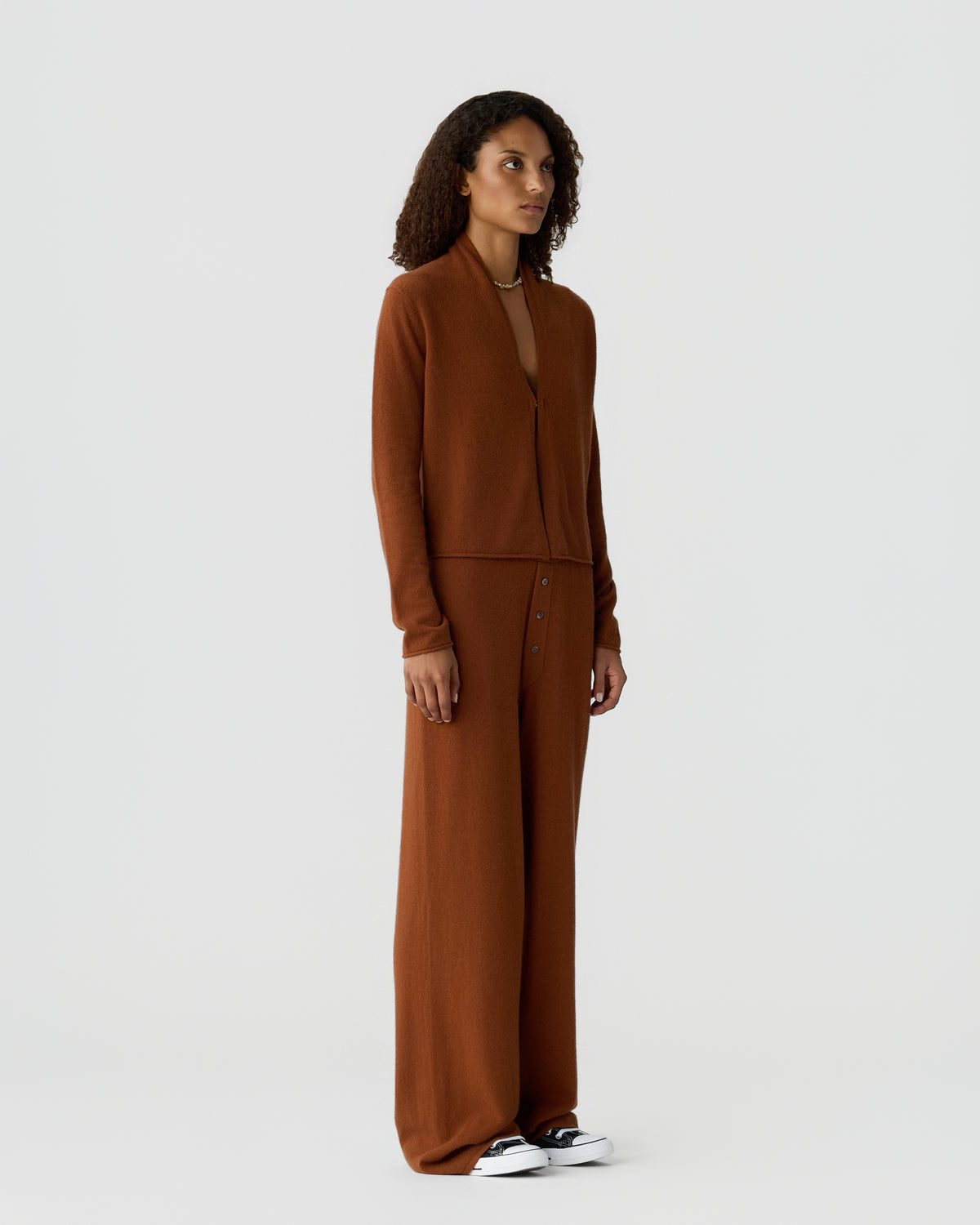 Woman wearing a monochromatic brown long sleeve knit top and wide leg trousers with white sneakers against a light gray background