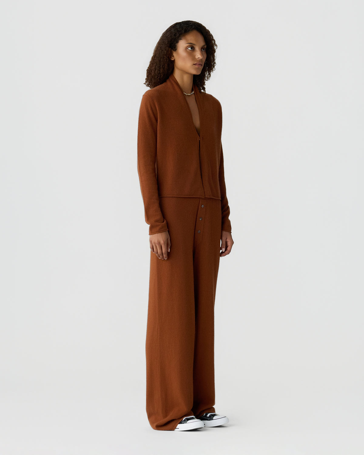 Woman wearing a brown cashmere cardigan and matching wide leg trousers with black and white sneakers against a light gray background.