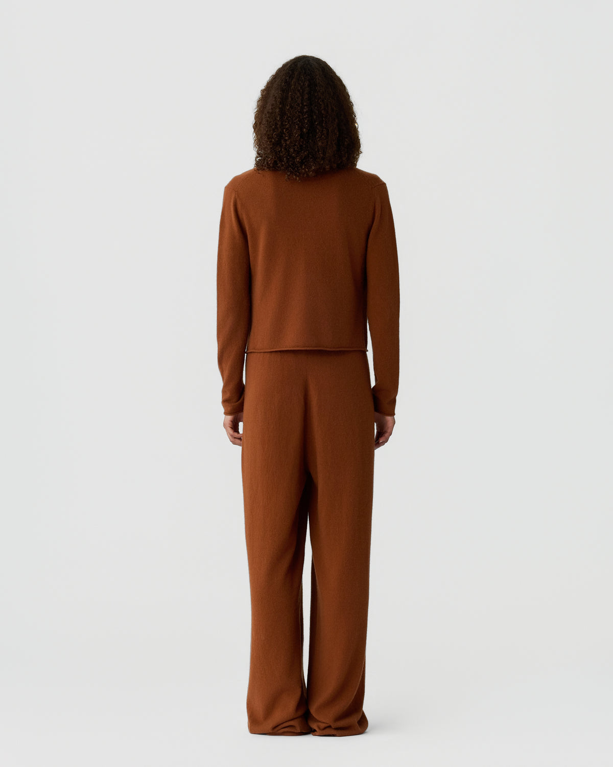 Back view of a person with curly hair wearing a brown long sleeve knit top and matching wide leg trousers against a plain light background