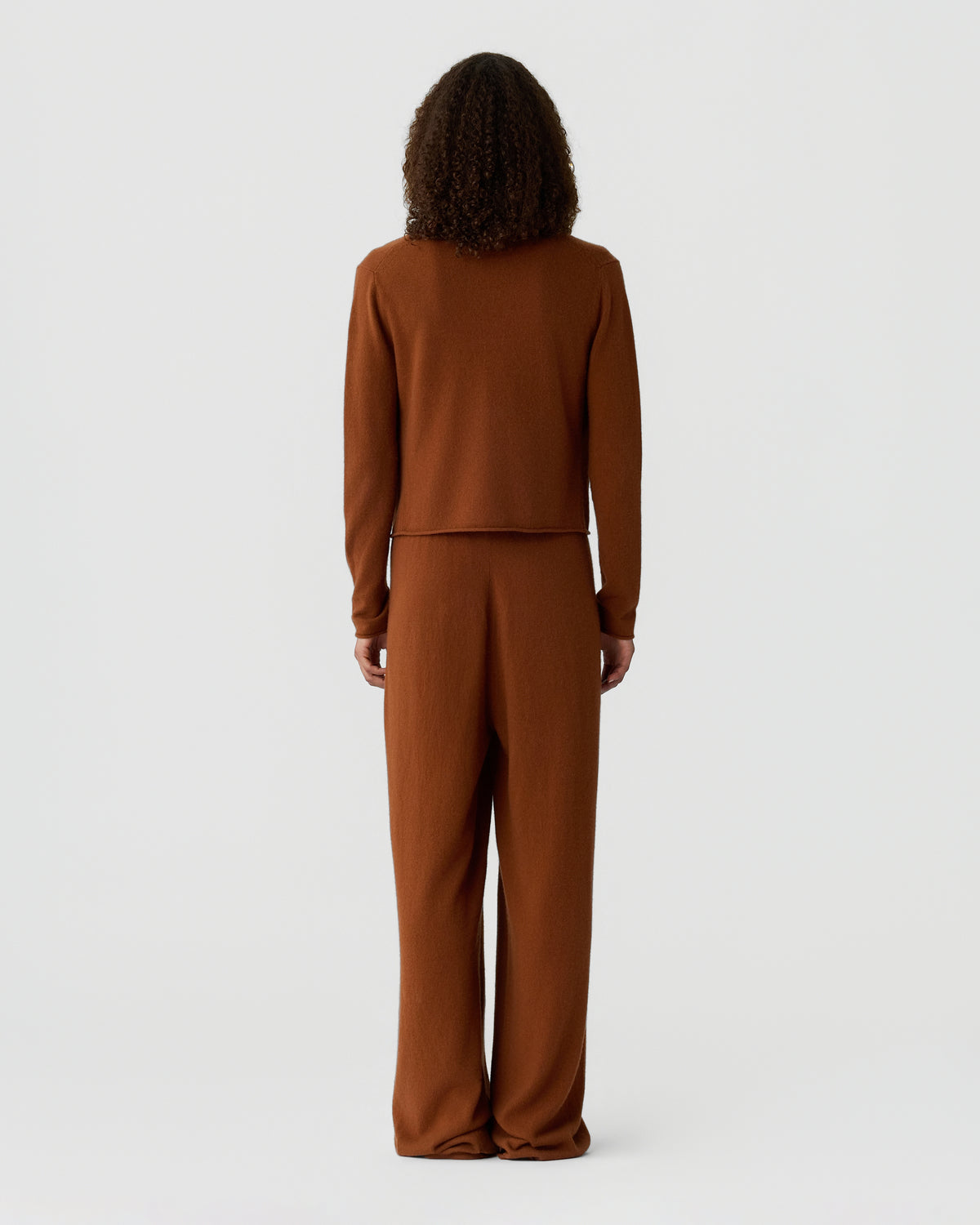 Rear view of a person with curly hair wearing a rust brown long sleeve top and matching wide leg trousers against a light gray background