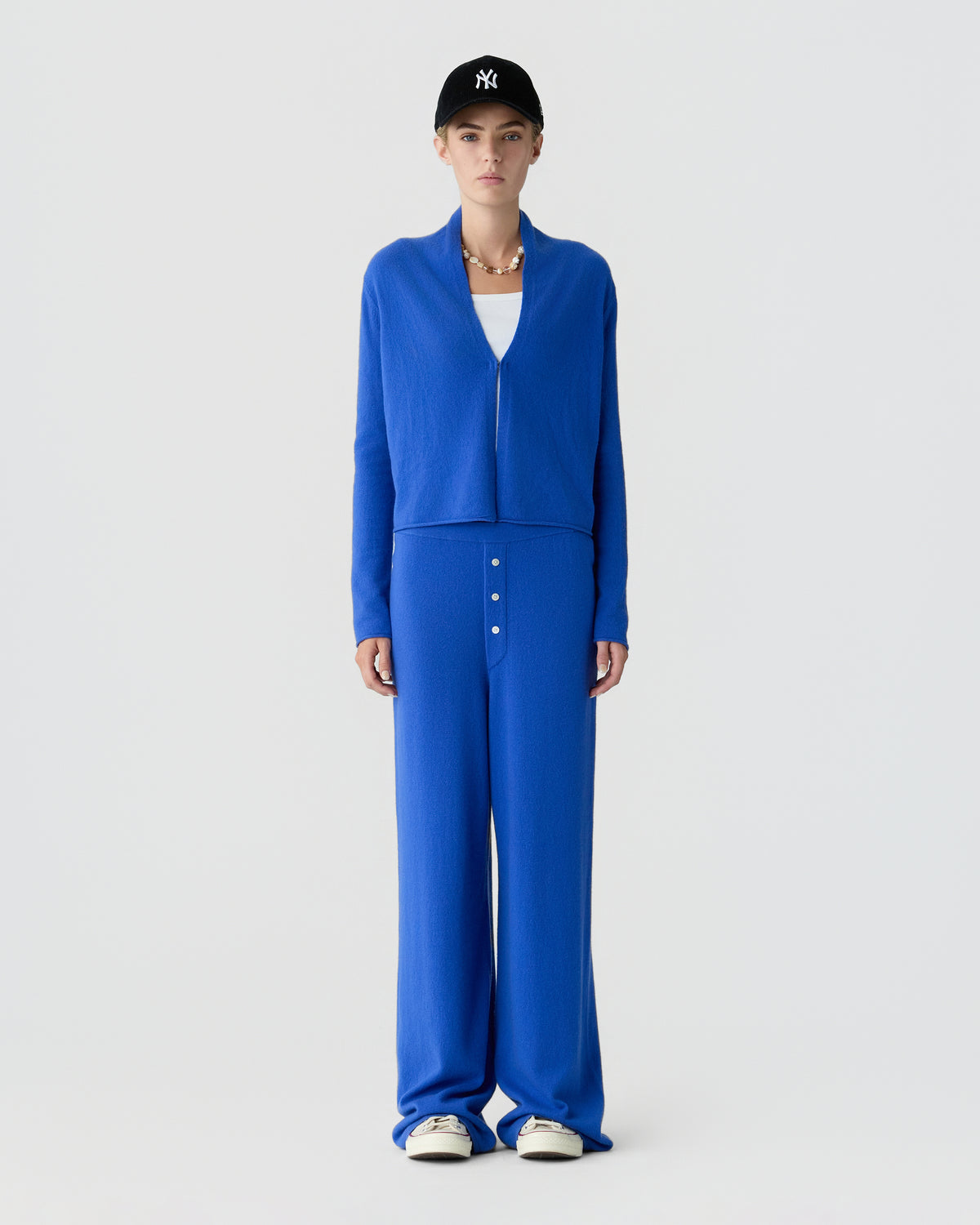 Model wearing a bright blue cashmere zip sweater and matching wide leg trousers with a black baseball cap and white sneakers against a gray background