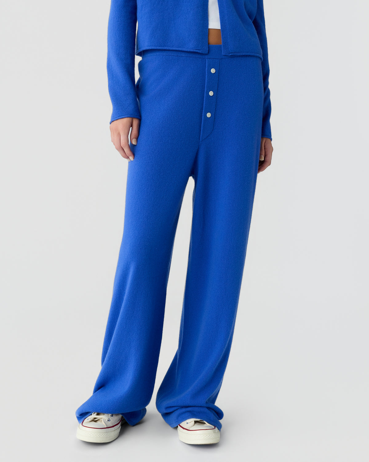 Cropped view of a person wearing a bright blue knit cardigan and wide leg trousers with button details, paired with white sneakers against a light background