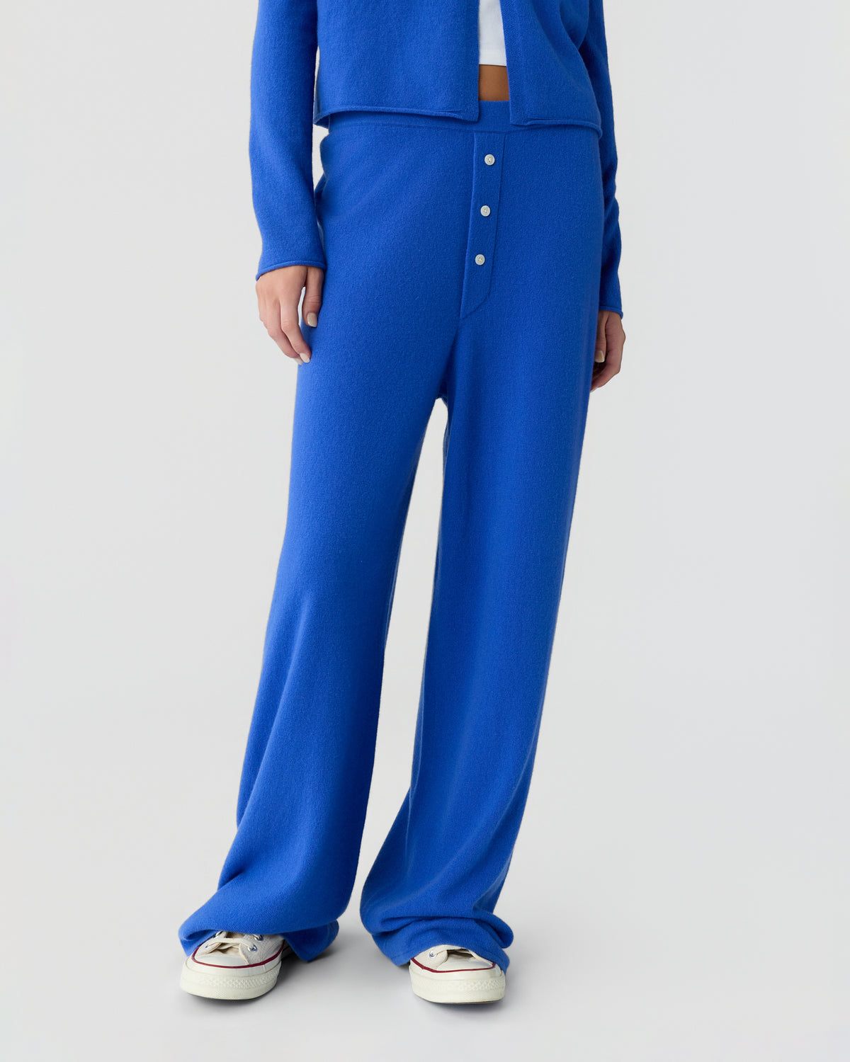 Cobalt blue wide leg knit trousers with a buttoned front, shown on a model wearing white sneakers against a light gray studio background.