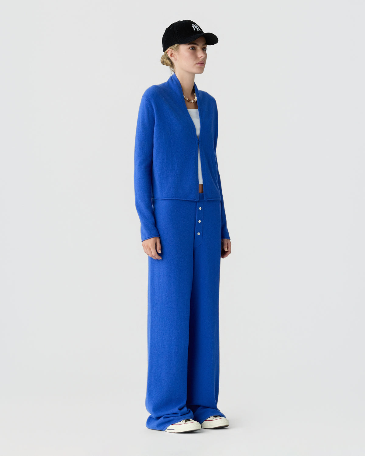 Woman wearing a bright blue cardigan and wide leg trousers with white sneakers and a black cap against a light gray studio background