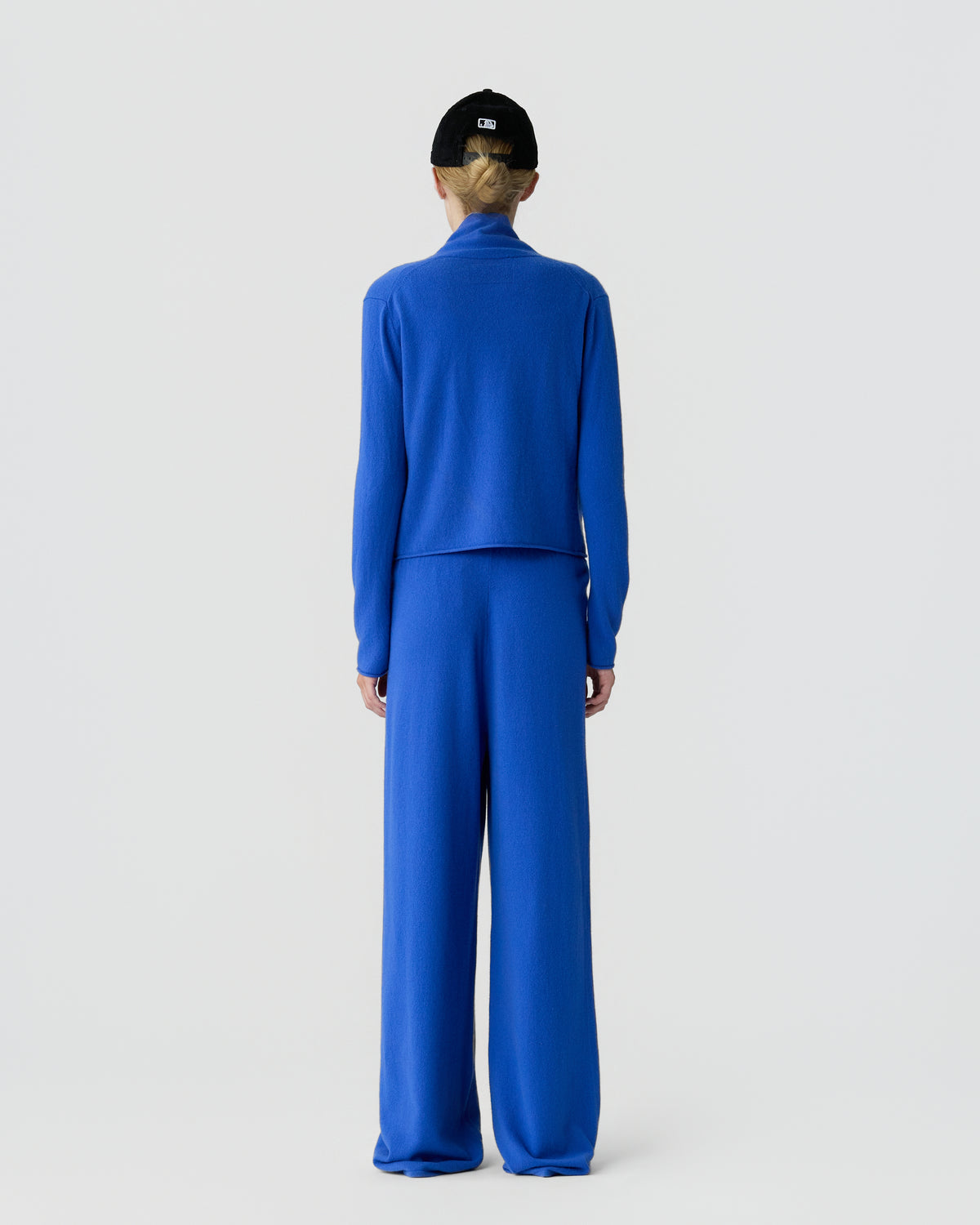Person viewed from behind wearing a bright blue long sleeve top and matching wide leg pants with a black cap against a light gray background