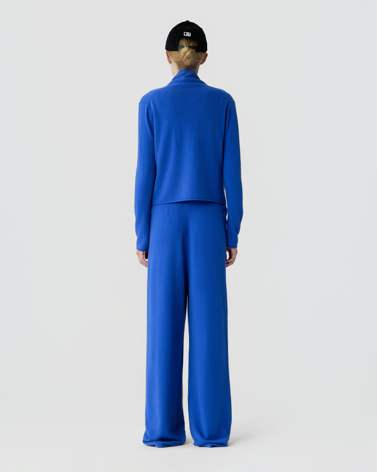 Back view of a model wearing a bright blue long sleeve top and wide leg trousers, standing against a plain light gray background.