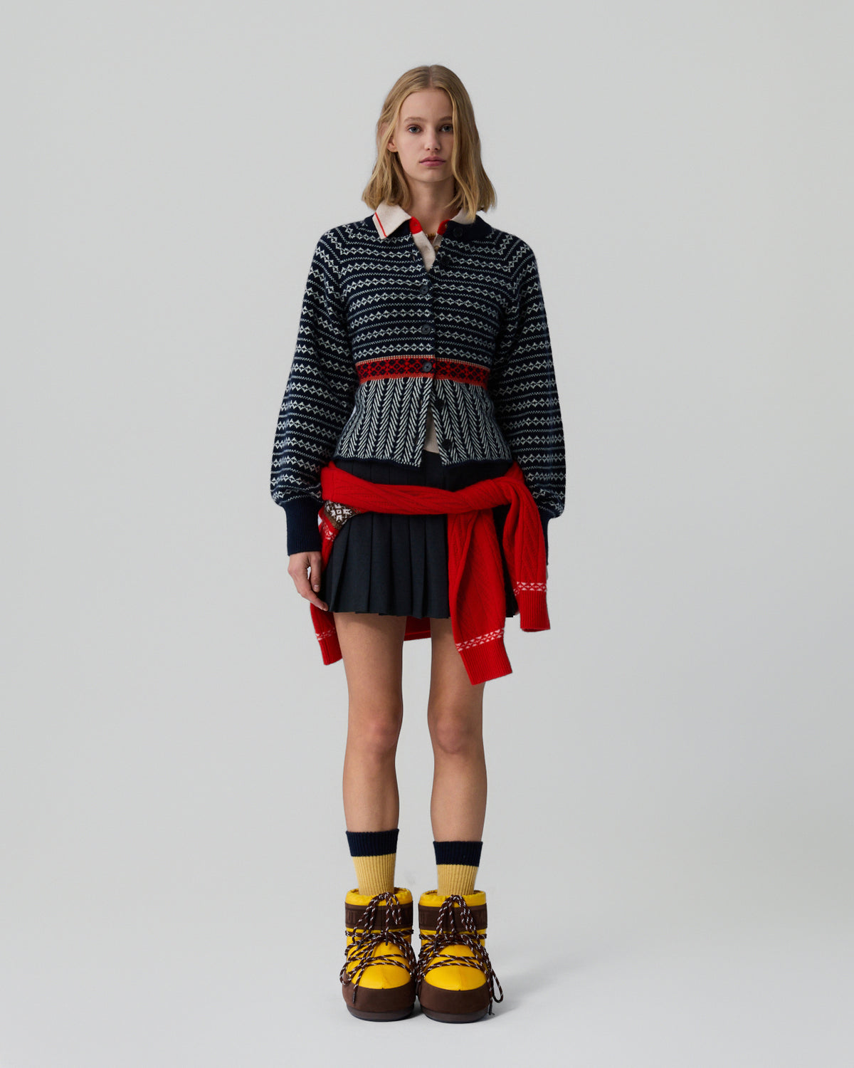 Young woman standing against a plain background wearing a navy patterned knit cardigan with a red waistband over a white collared shirt, paired with a black pleated skirt, a red sweater tied around the waist, yellow and black socks, and yellow lace-up boots.